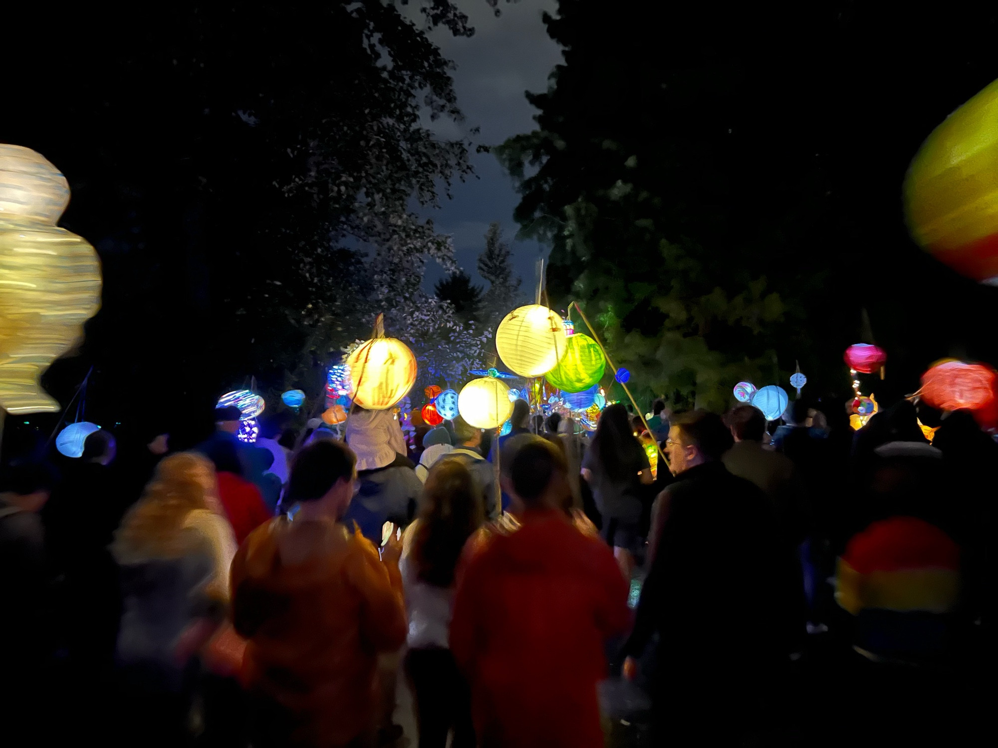 A crowd walking down a park path at night with multicolored lantens