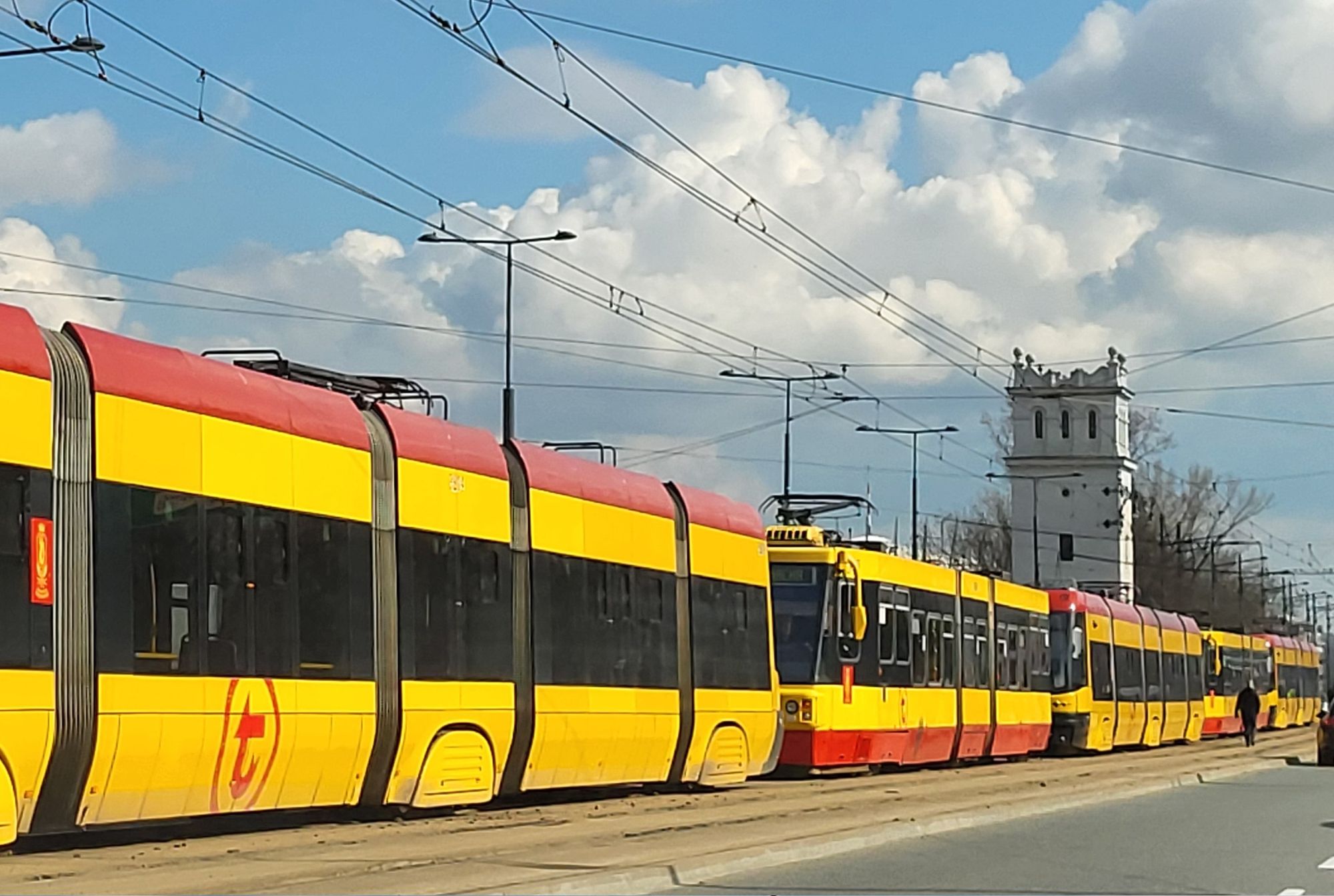 Eine Aneinanderreigung von Straßenbahen mit gelb/ roter Färbung, aber unterschiedlicher Typen. Oben blauer Himmel mit Oberleitungen.