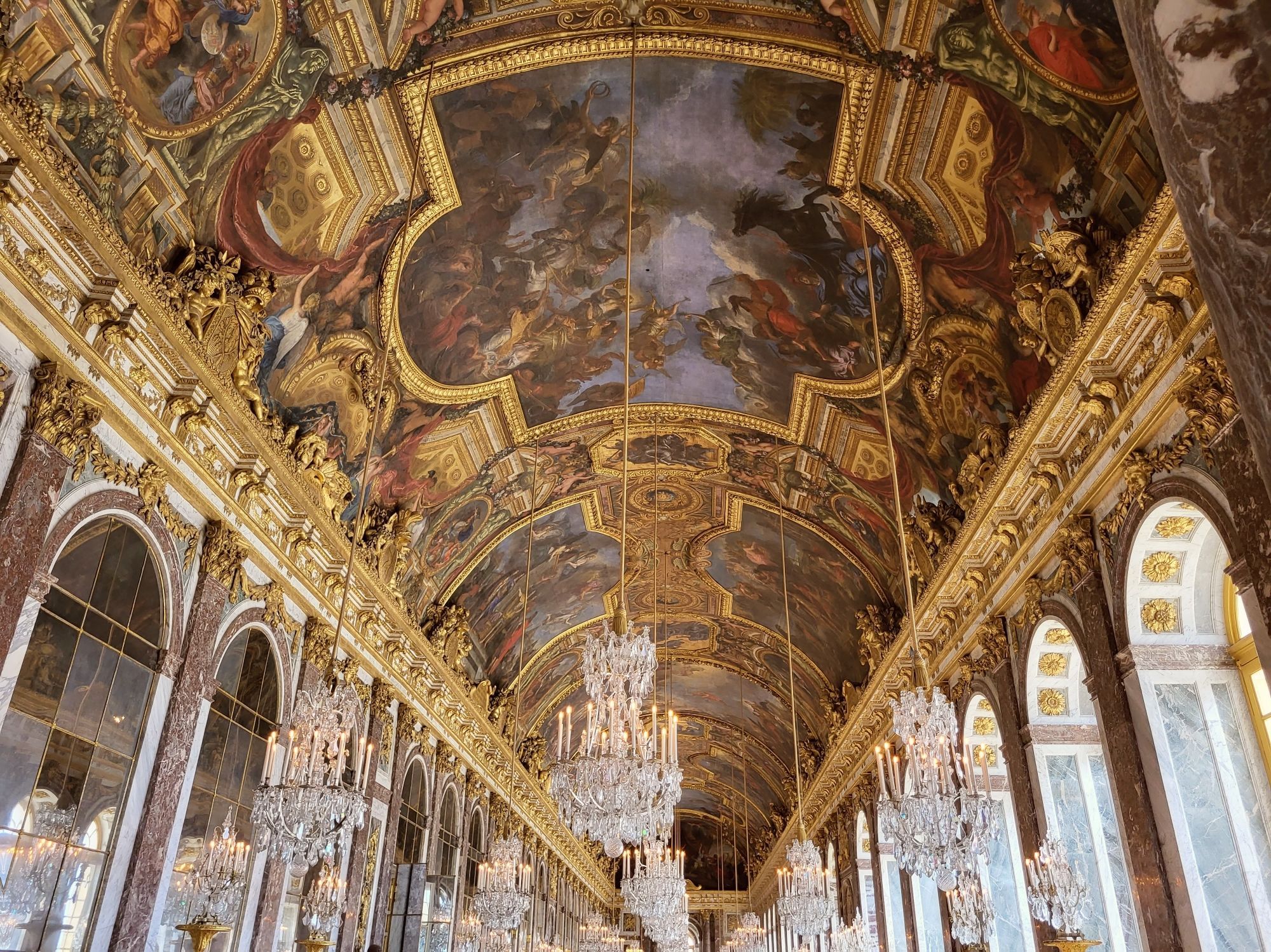 Eine prächtig barocke bemalte Decke mit vielen goldenen Verzierungen. Darunter hängen kristallene Kronleuchter. Links sind Spiegelflächen erkennbar. Rechts in gleicher Höhe Rundbogenfenster.