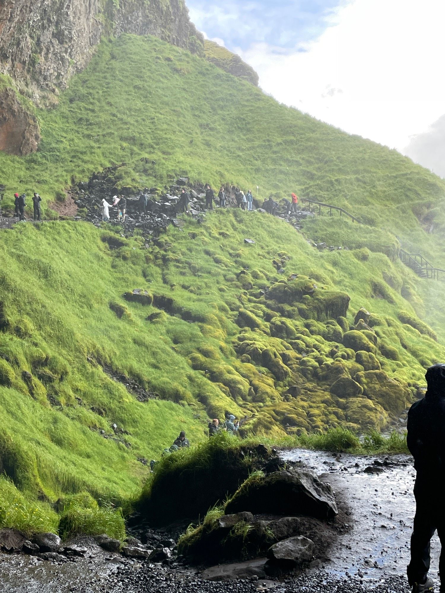 People on the trail approach to go behind the falls. The stairs are barely visible amid the moss on the right. The photo is taken from a much lower height behind the falls.