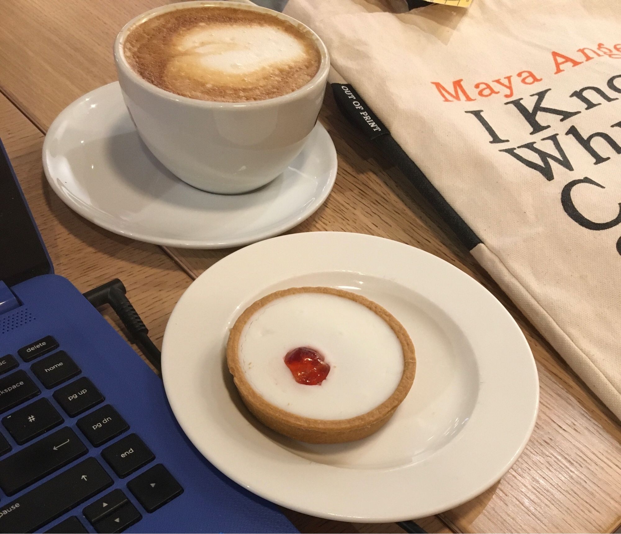 Laptop, coffee, Bakewell tart, and Maya Angelou tote bag on cafe table