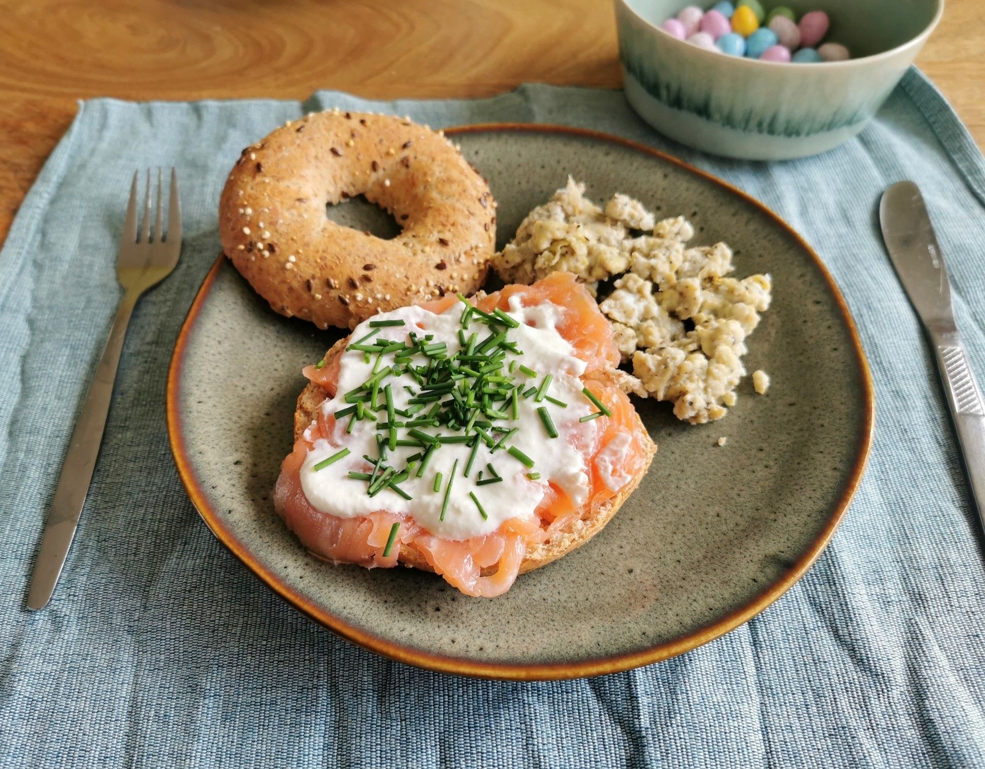 Een close up van een grijsgroen aardewerken bordje met daarop een meergranen bagel belegd met gerookte zalm, mierikswortelsaus en bieslook met ernaast roerei met kruiden. Boven in de hoek staat een schaaltje met pastelkleurige paaseitjes.