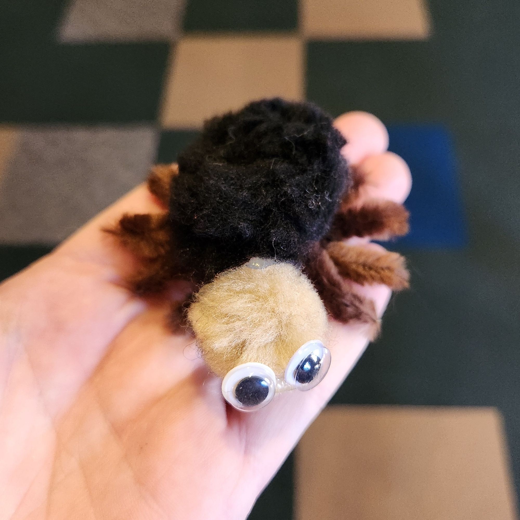 The same creature from above, made of black and tan pompoms and googly eyes. Now it is lying down and clearly has 8 tiny legs made of brown pipe cleaners