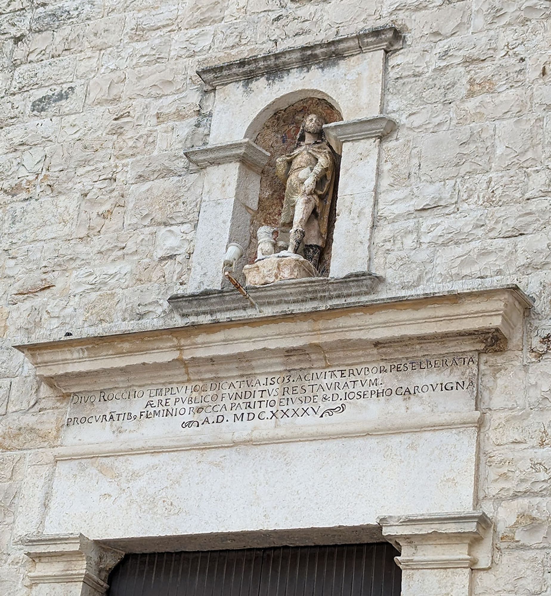 The inscription of 1645 is on the architrave of the simple portal of the white limestone building. A statue of San Rocco is in the niche above.