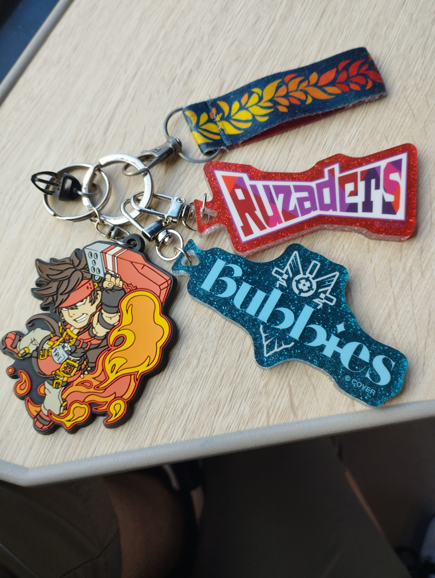 A detachable phone charm with four charms on it, one showing the character Sol Badguy in his Guilty Gear Xrd appearance in the middle of the Volcanic Viper special, two glitter charms, both featuring the fan names of the Holostars VTubers Goldbullet and Crimzon Ruze, Bubbies and Ruzaders respectively, in designs similar to their logos and a small wrist strap with a laurel design going from yellow to red in the style of the HADES loading screen.