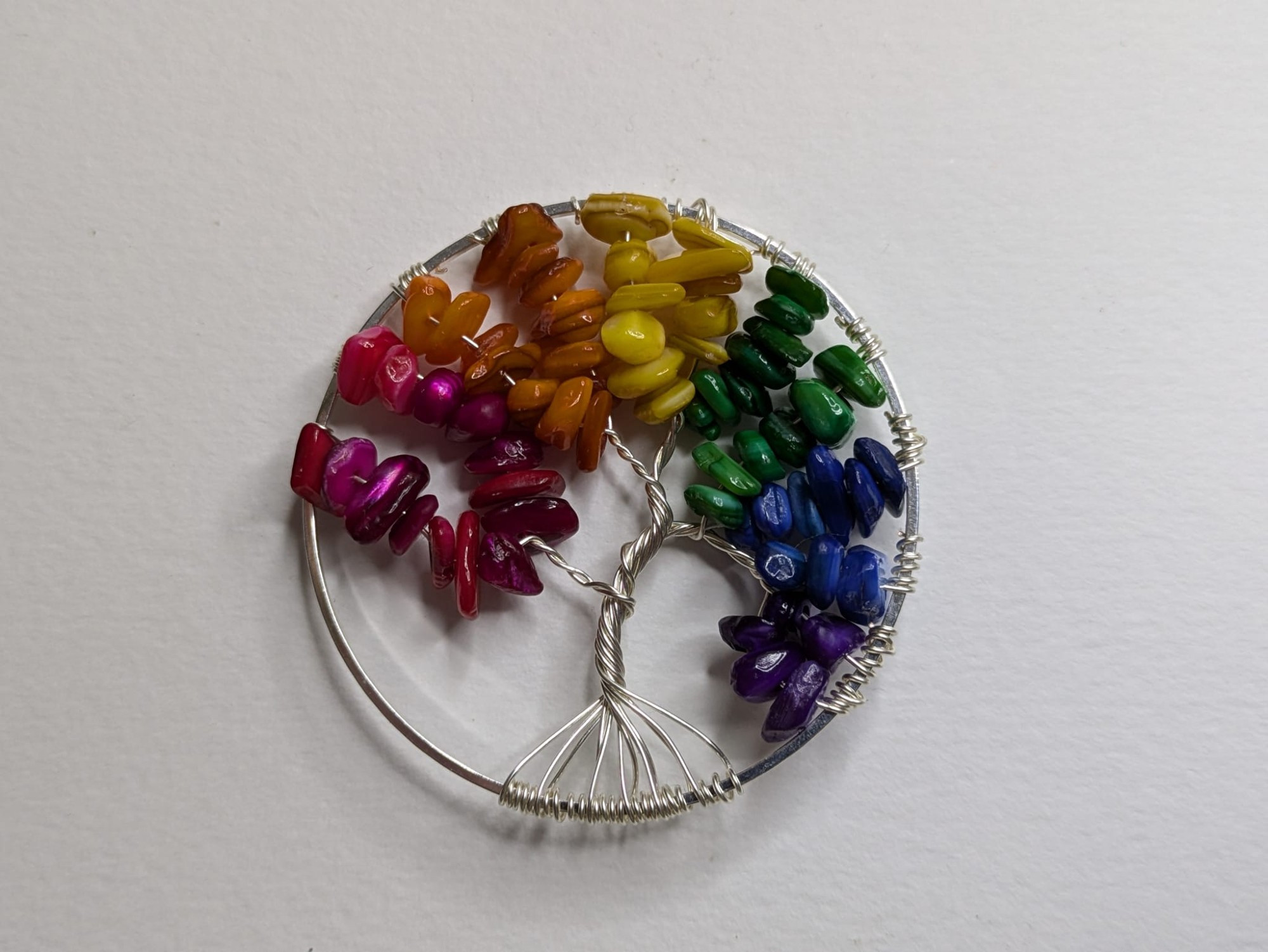 A round pendant sits on a white background. The pendant is a silver circle filled with a wire tree. The tree has leaves composed of bits of shell in the colors of the rainbow, from left: Red, orange, yellow, green, blue, and purple. There are two wire branches of each color.