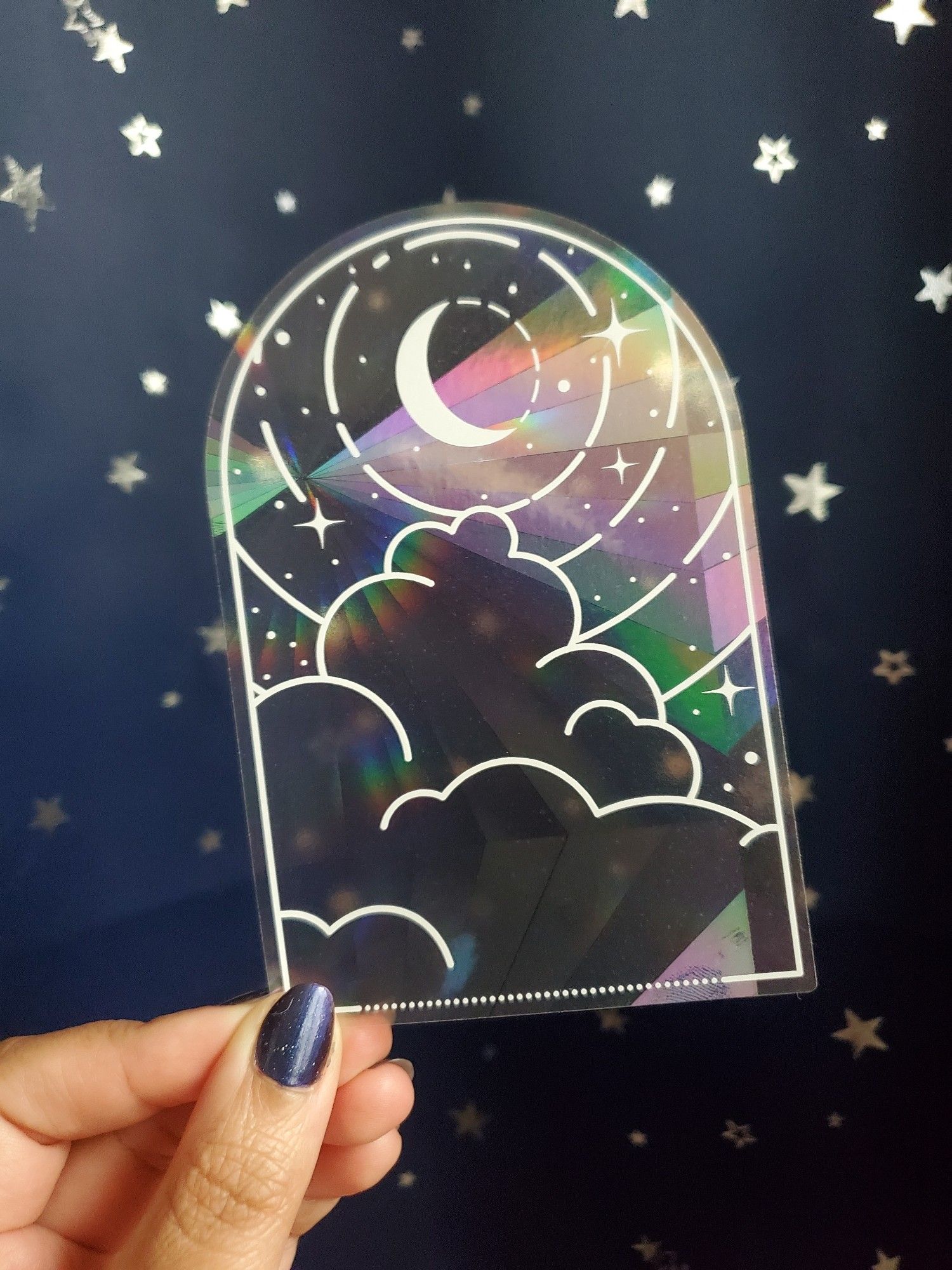 Photo of the sun catcher sticker heals by the artist in front of a dark blue background with silver stars. It's clear with white print od clouds, crescent moon, concentric circles, and twinkling stars. You can see the radial axicon pattern that creates rainbows.