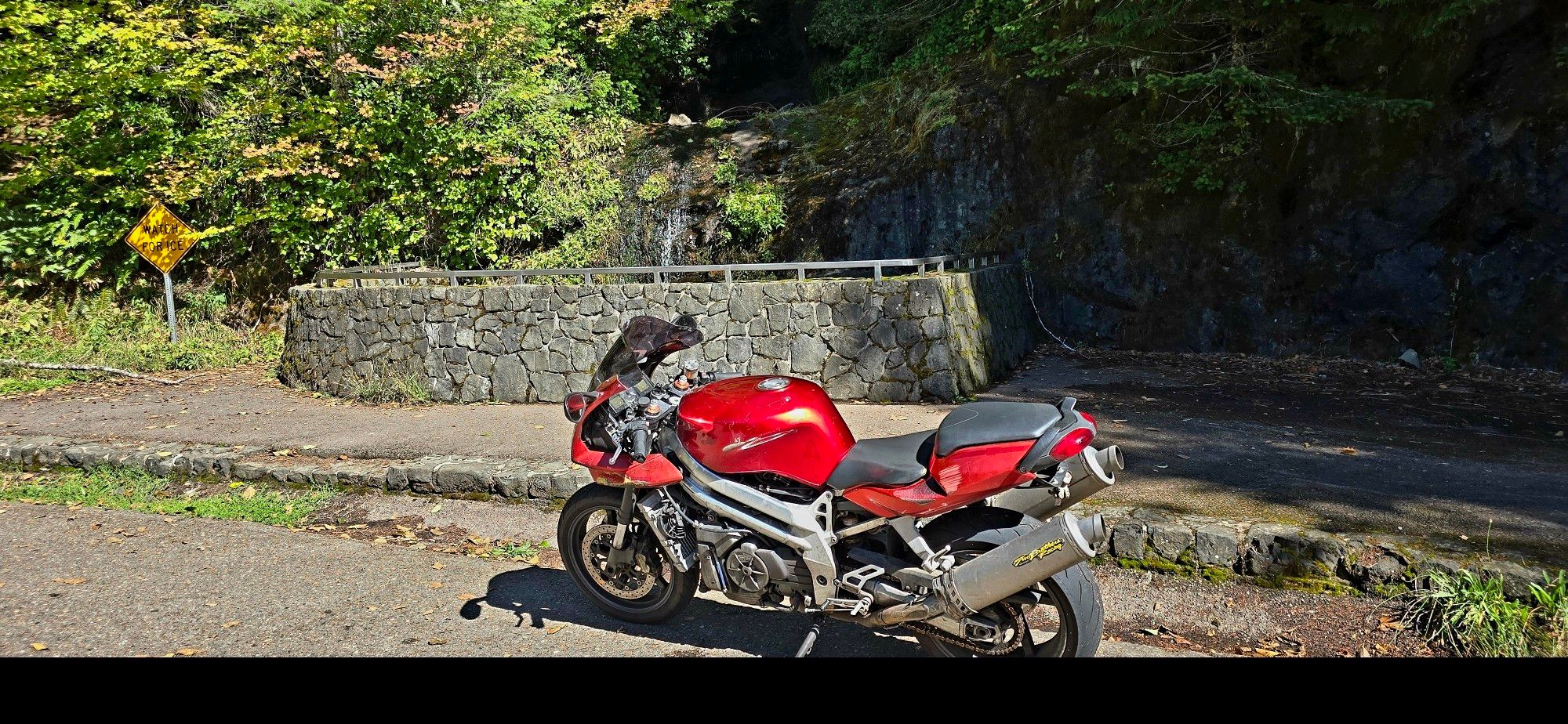 Red aprilia sl1000 falco parked on the side of the road infront of a forested area with a waterfall