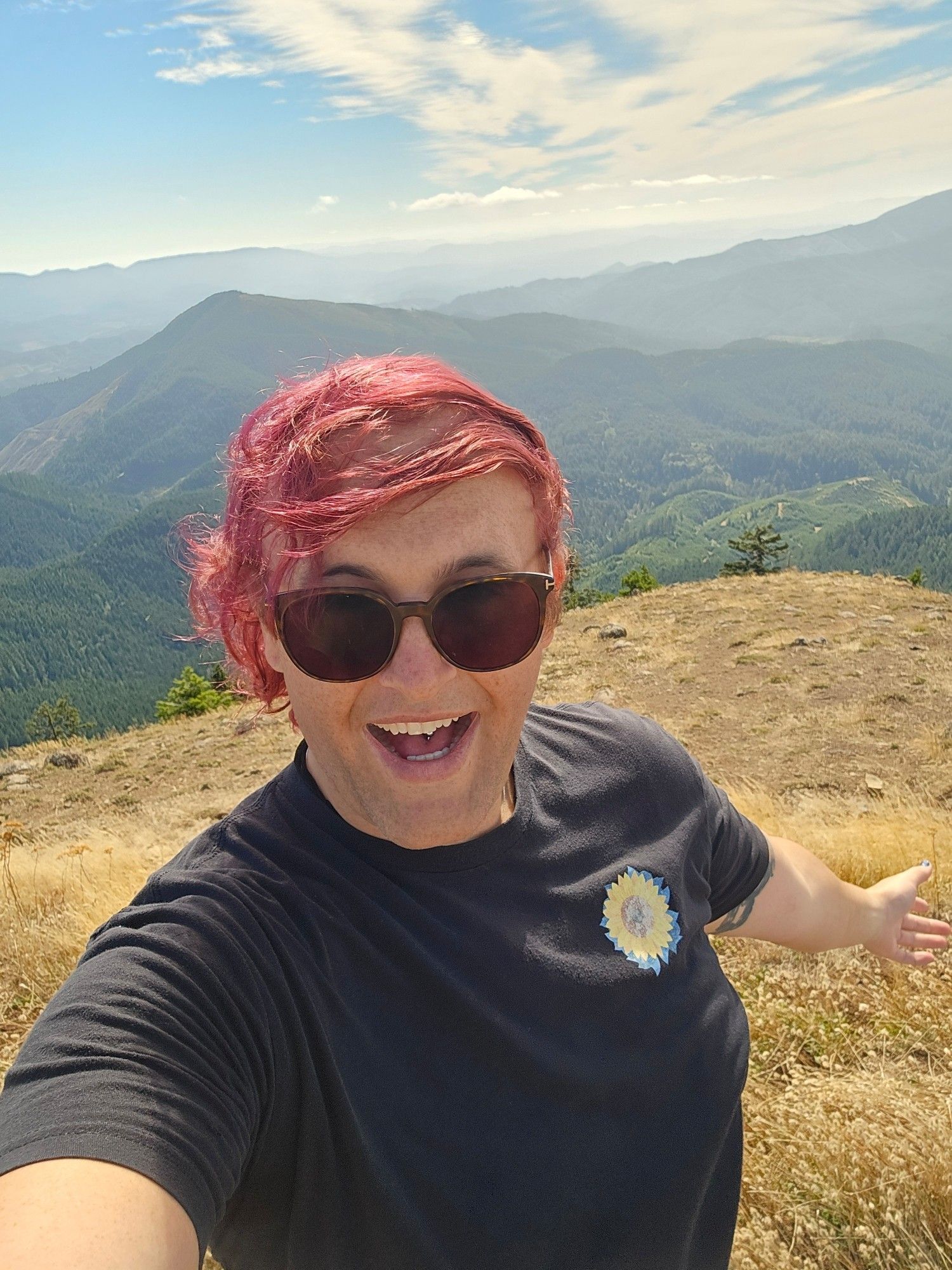 Lila standing on top of Mary's Peak woth a big smile, her glasses, and a sunflower badged shirt. Her purple hair glowing in the sunlight