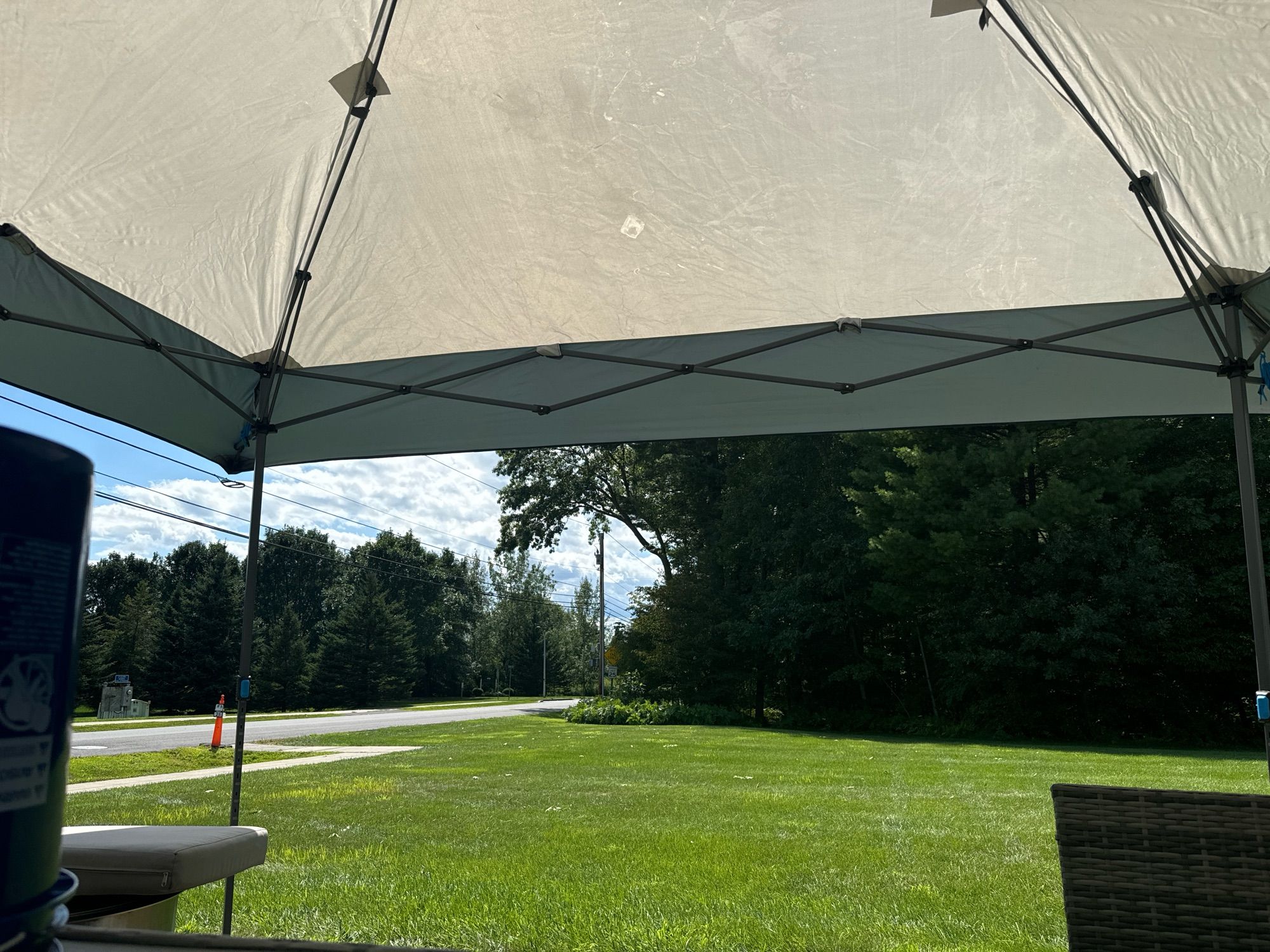 Green grass yard with trees/ woods behind. Our from under the sun shade canopy you see blue sky and clouds above the trees.