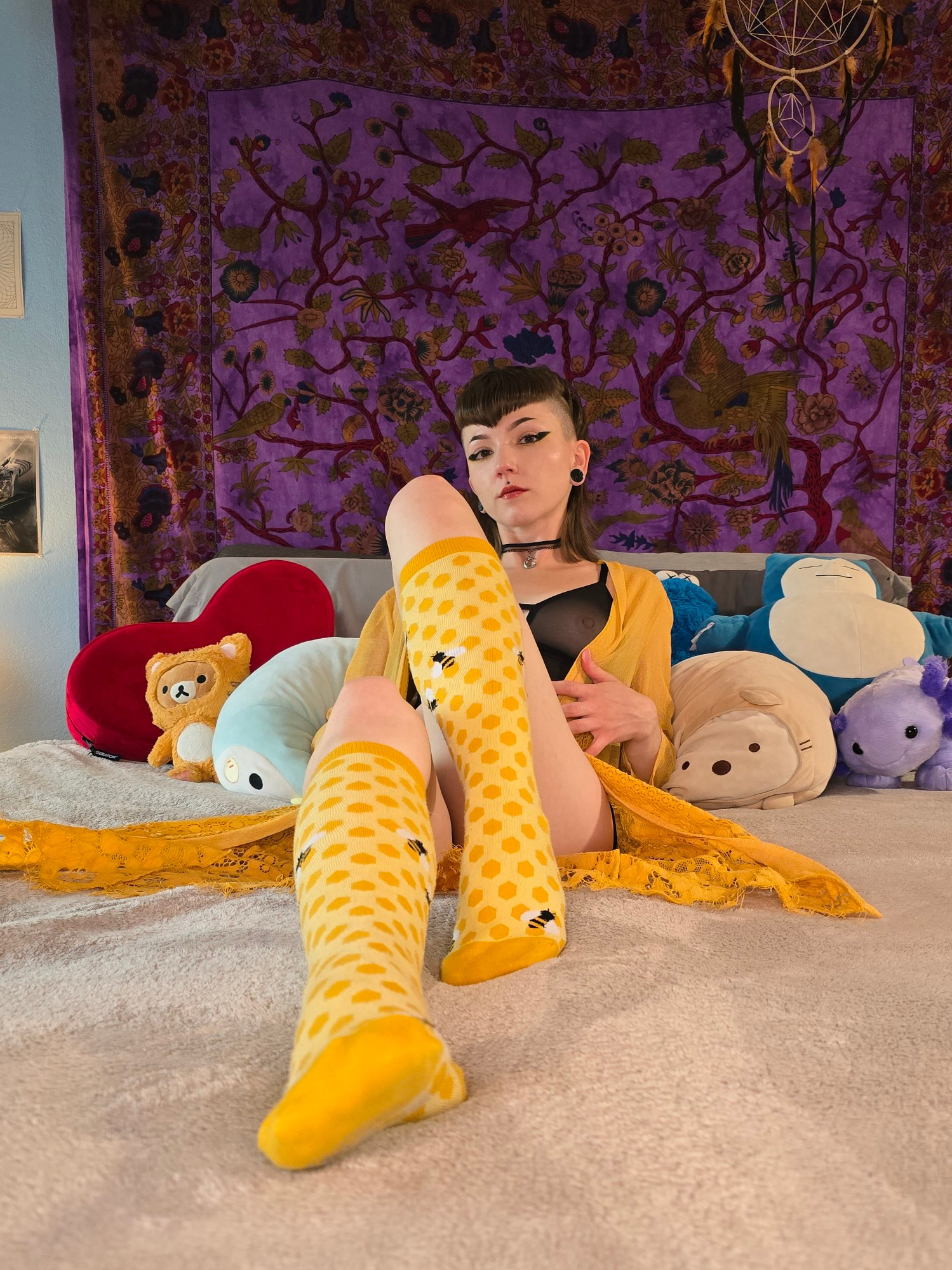 Honey is laying in her bed, propped up on her elbows. She is wearing a bright yellow lace cardigan and knee high socks with a yellow honeycomb pattern, and a black lace choker. There is a dark purple tapestry pinned to the wall behind her, and she is surrounded by her stuffed animals. Among them, snorlax, a purple axolotl, and Kuma bear in a cat suit. 