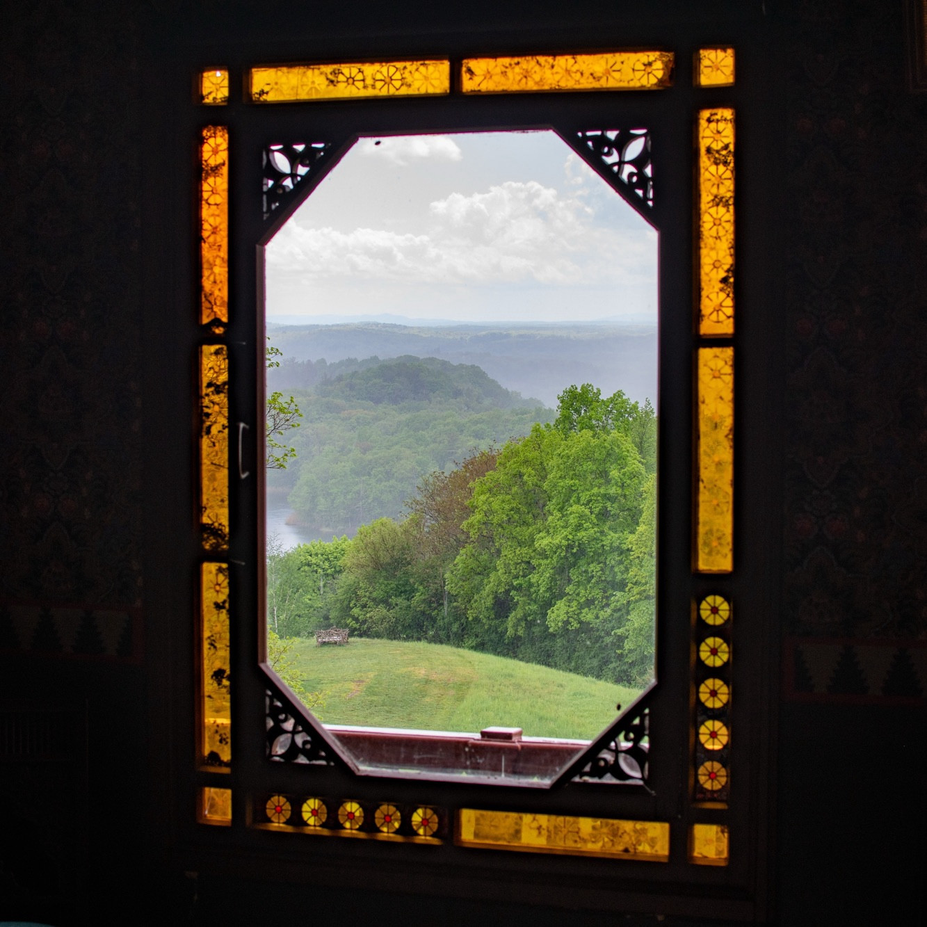 The view out of a window with a stained glass border. Outside, you can see lush trees and rolling green hills with mist. The border and dark walls give the illusion that the window is actually a painting.