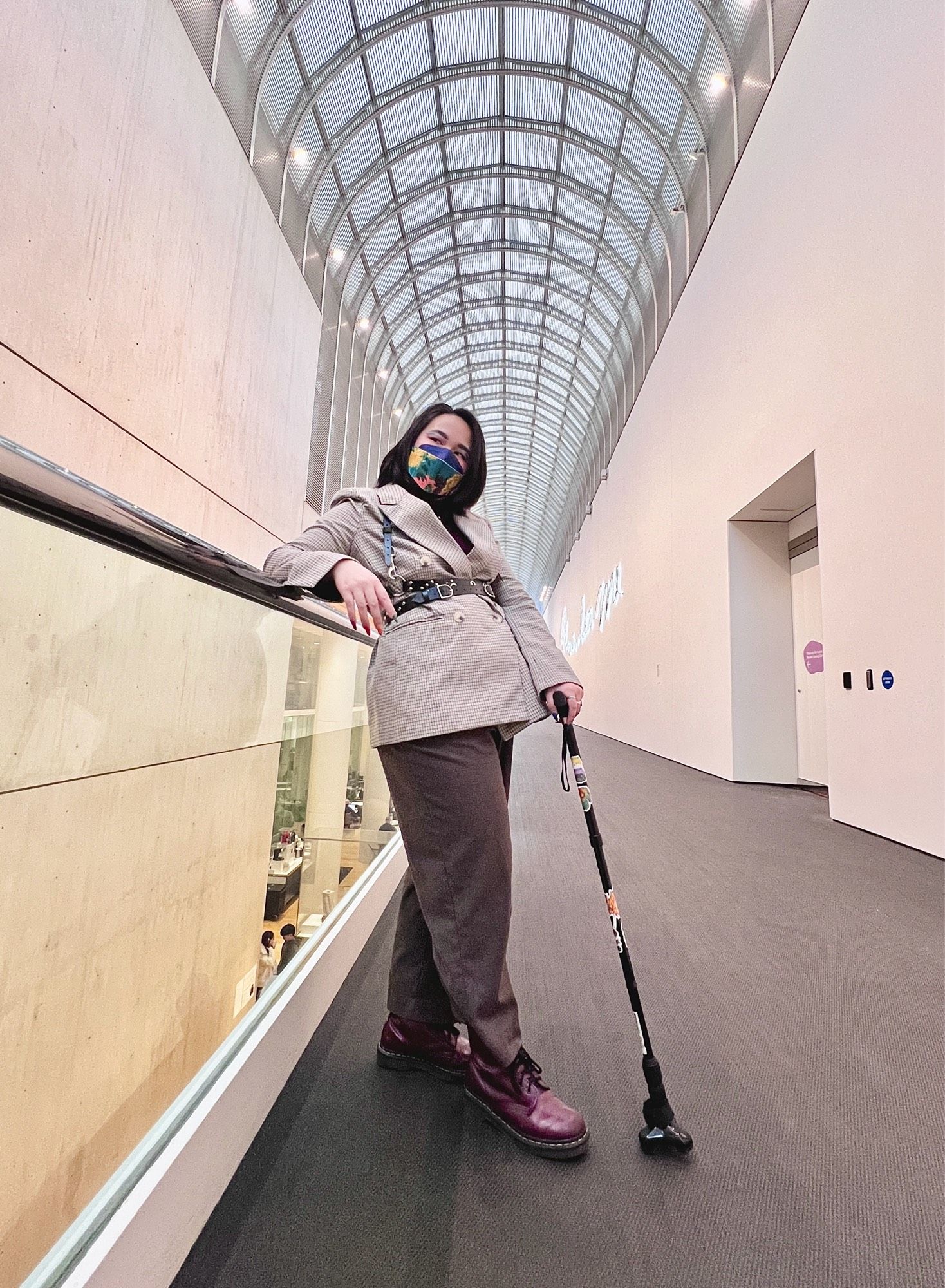 Jae stands in the museum of fine arts, dressed in a blazer with a harness worn over. They lean against the railing, cane in other hand.