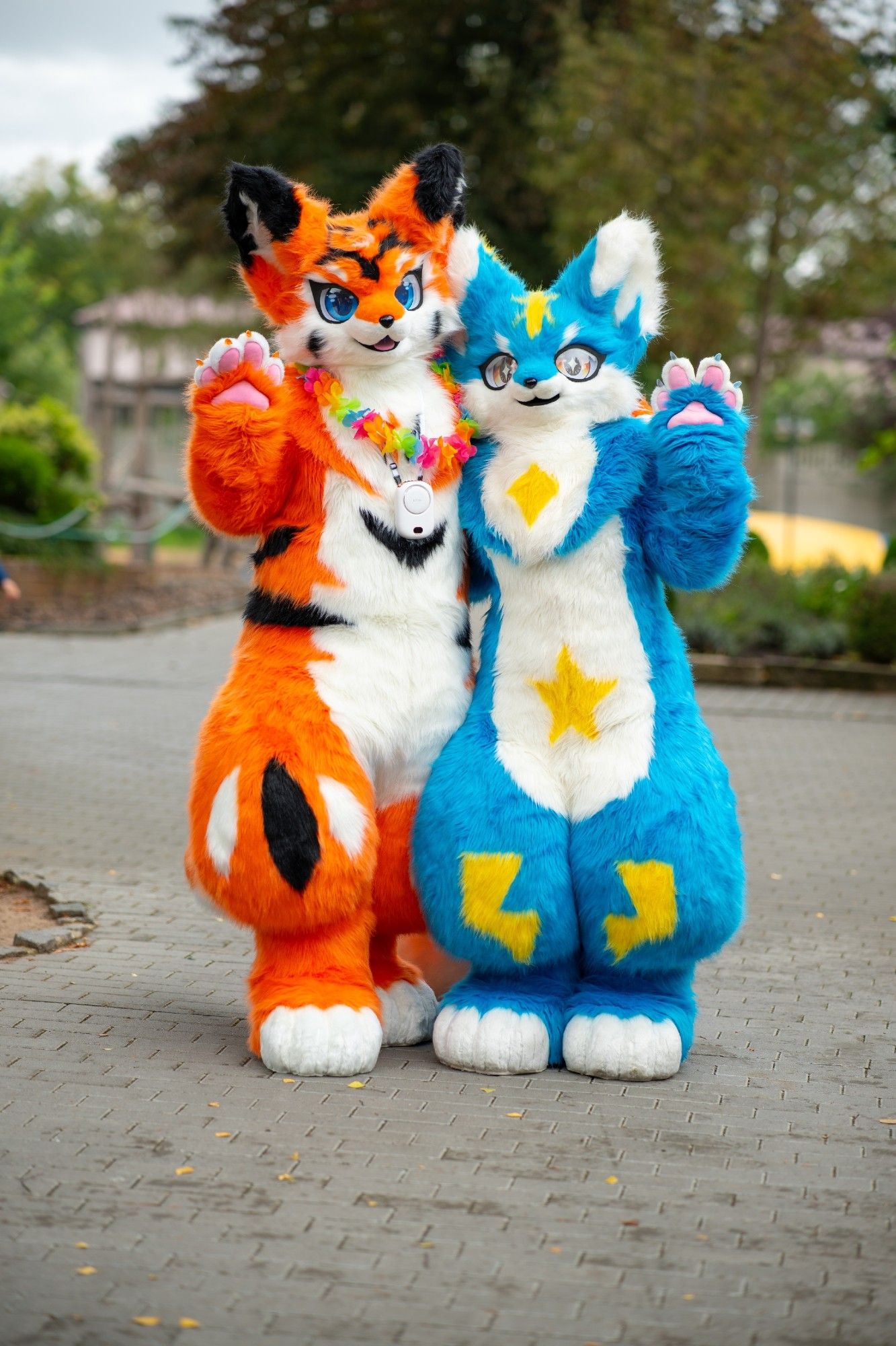 An orange fox with blue eyes and black markings and a blue cat with yellow eyes and yellow markings standing together waving.