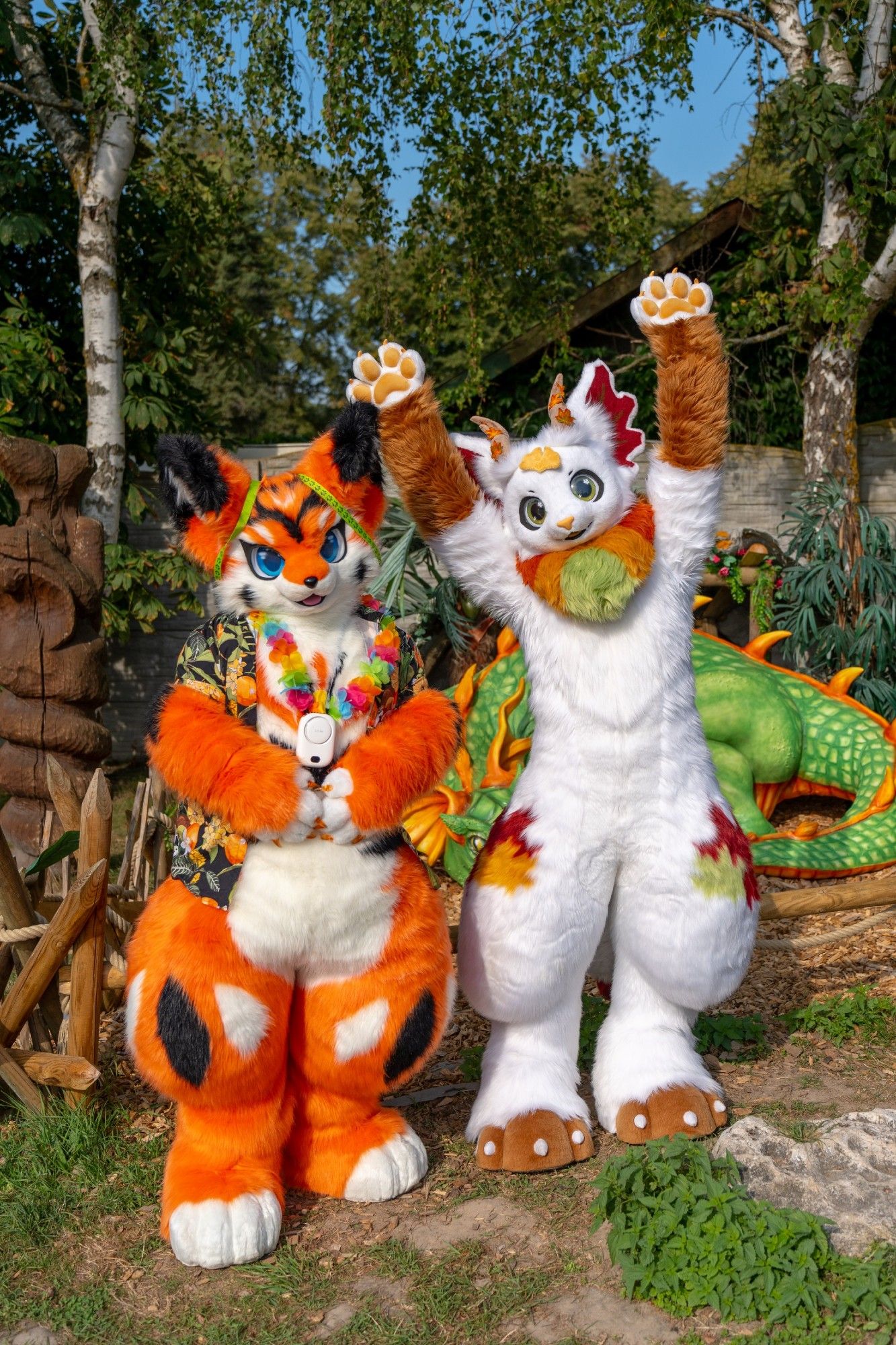 An orange fox with blue eyes, black and white markings, a Hawaiian shirt with a floral pattern and a Hawaiian lei stands right next to a white-furred dragon with green eyes and colorful autumn patterns on its body, waving eagerly at the viewer. They are standing in front of a green dragon lying on the ground.