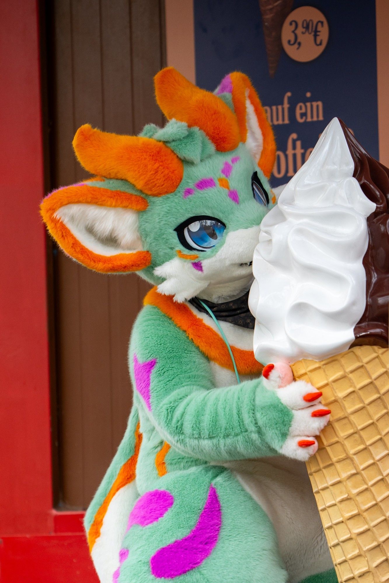 A green dragon with blue eyes and orange horns is licking on a large ice cream cone to cool off on a hot day