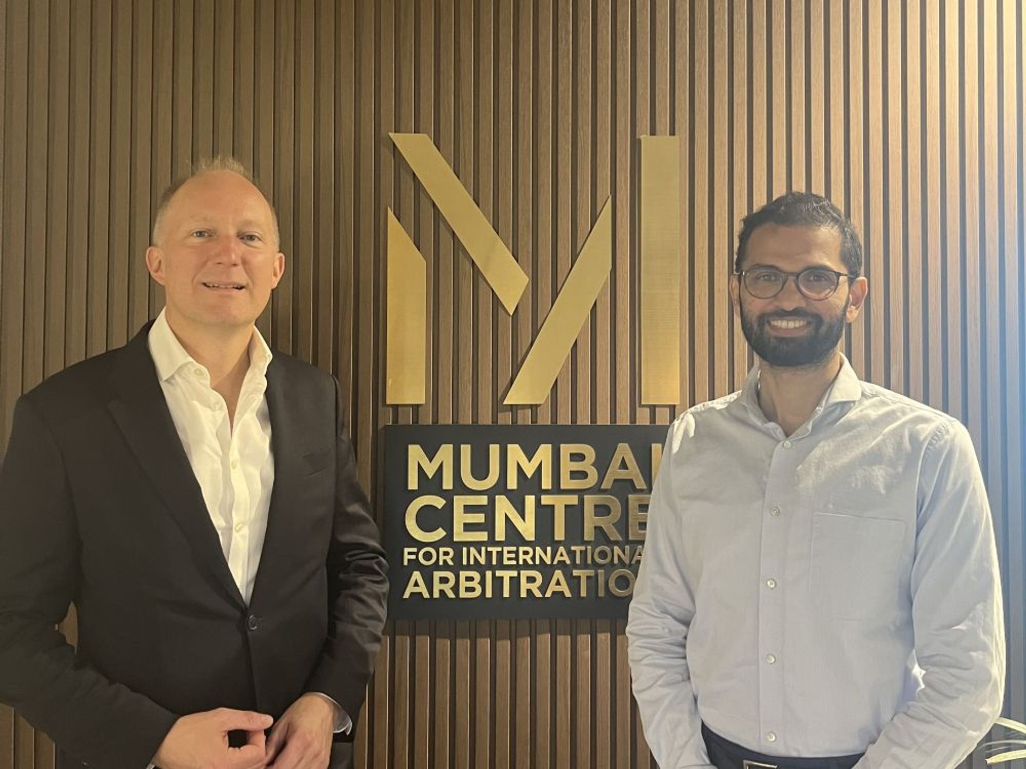 Sam Townend KC and Madhukeshwar Desai standing in front of a large logo for the Mumbai Centre for International Arbitration.