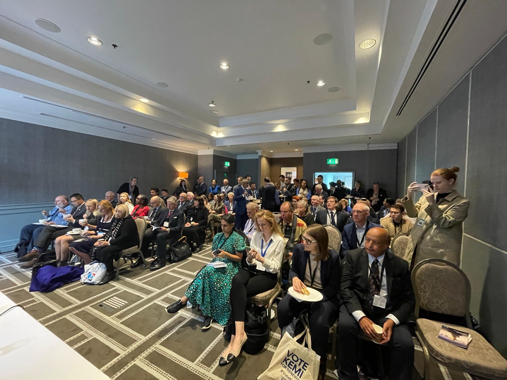 A conference room full of people sitting in rows of seats with some people standing at the back.