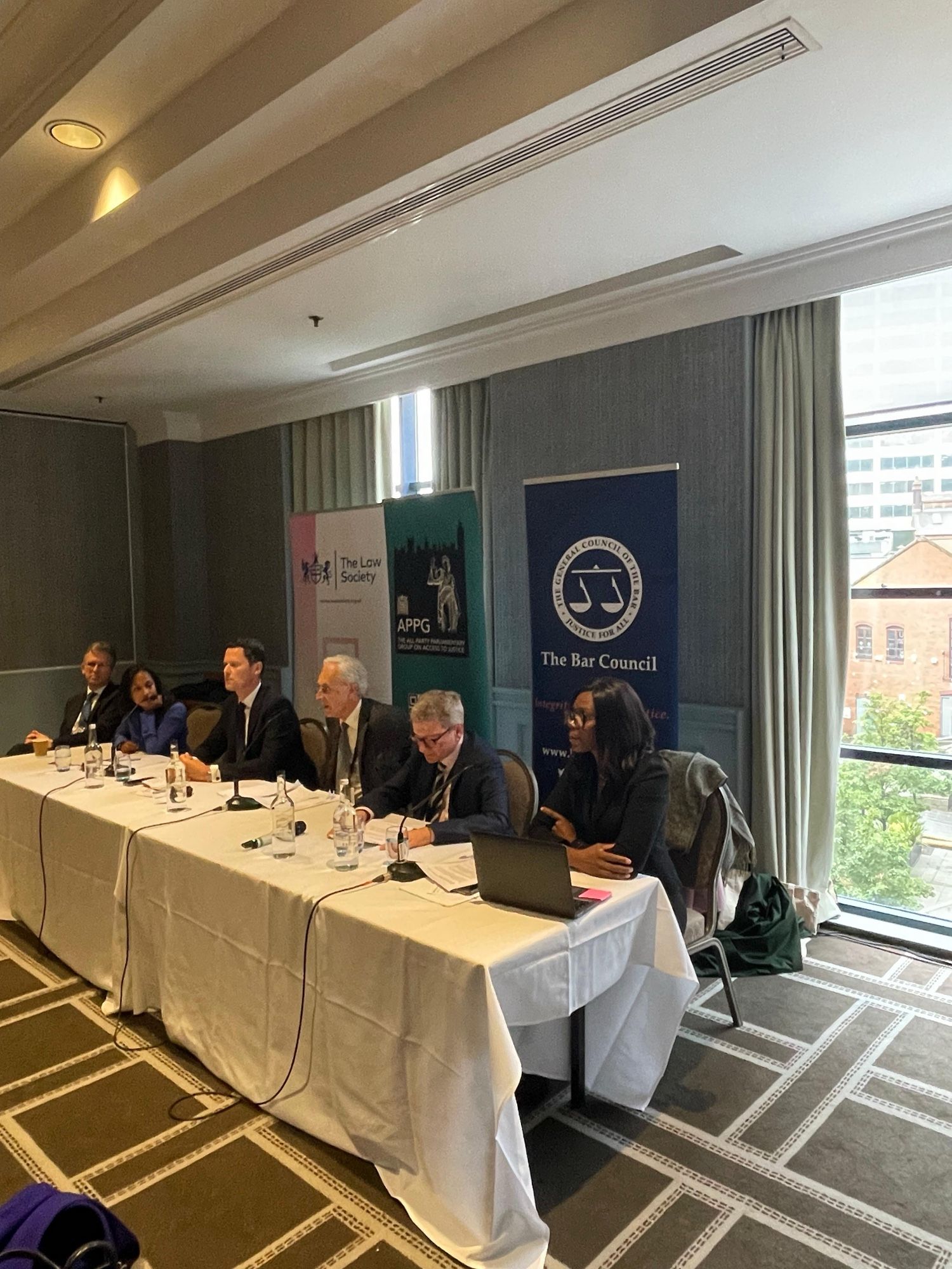 Long table of panel speakers with the banners of the Bar Council, the Law Society and the All Party Parliamentary Group on Legal Aid behind them.