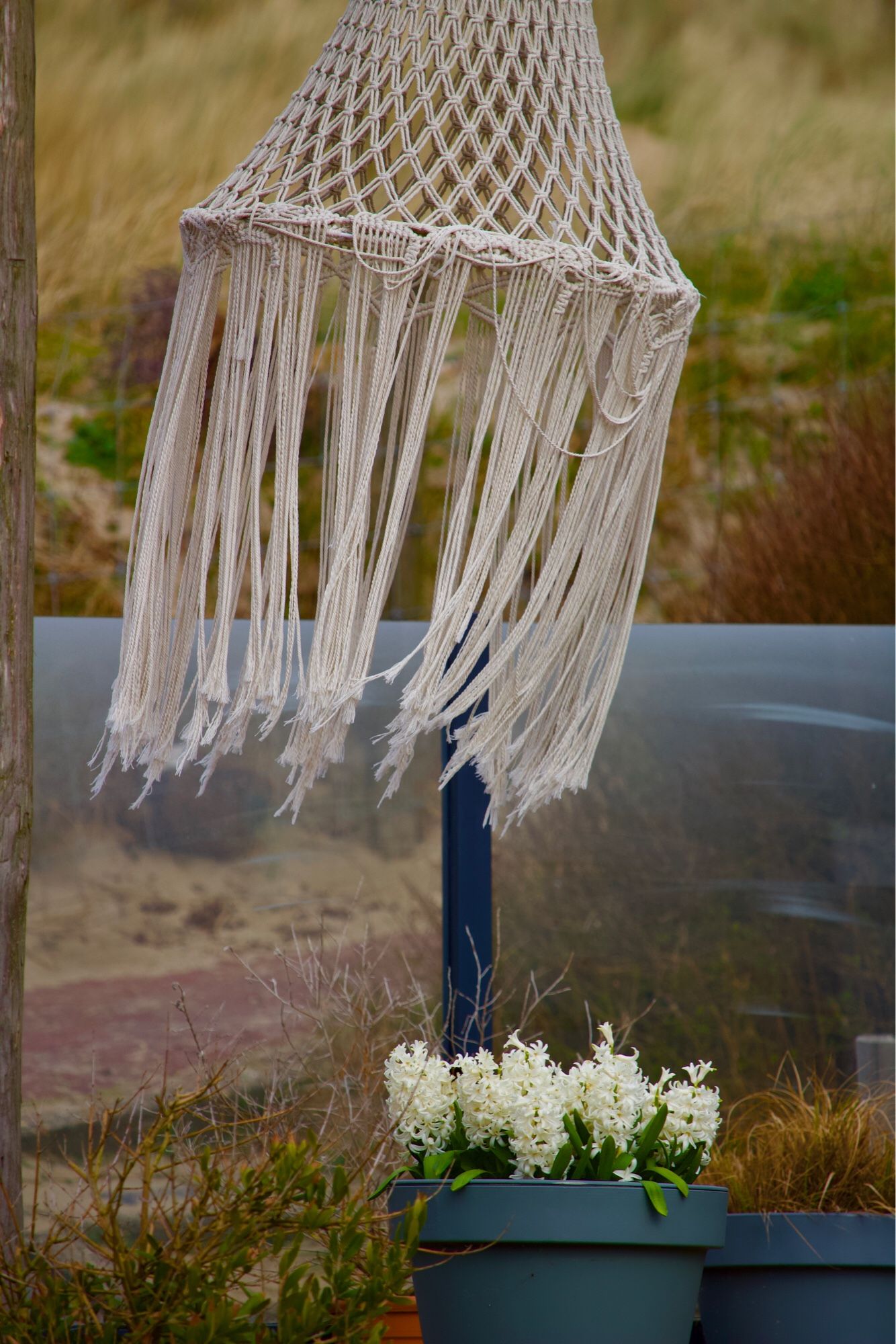 Een pot met witte chrysanten, daarboven hangt een grote katoenen, geknoopte lampion in de wind.