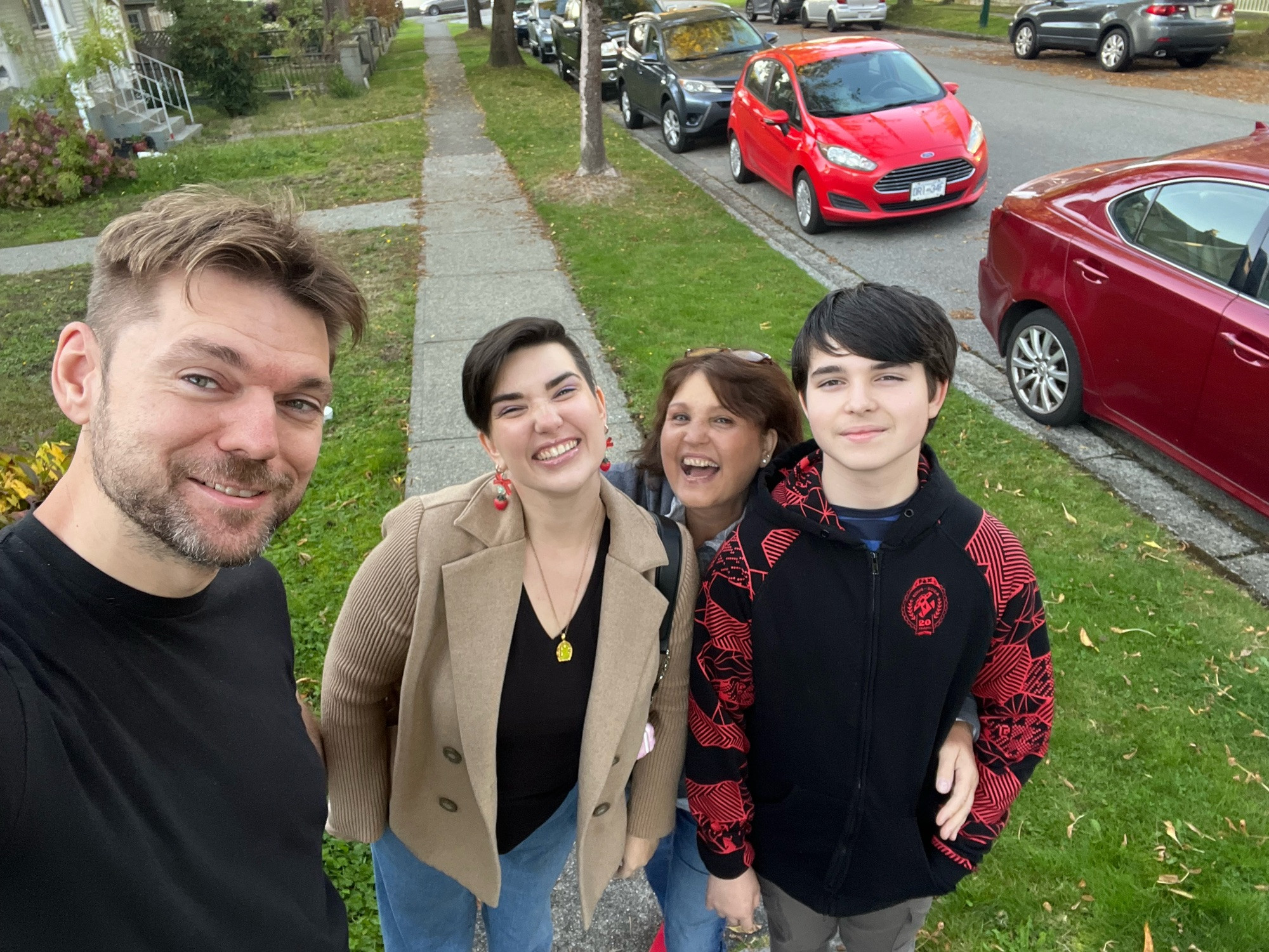 Four smiling people on a car lined street. Taller blonde man in a blue shirt holds the phone. Two women with dark brown hair grin (one in a camel coloured coat) and a young man in a hoodie smirks.