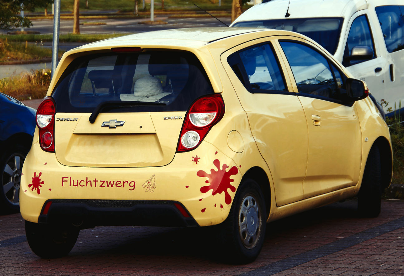 Foto eines gelben Kleinwagen der Marke Chevrolet. Auf dem hinteren Stoßfänger sind links und rechts jeweils ein Klecksartiger roter Aufkleber. In der Mitte steht 'Fluchtzwerg'