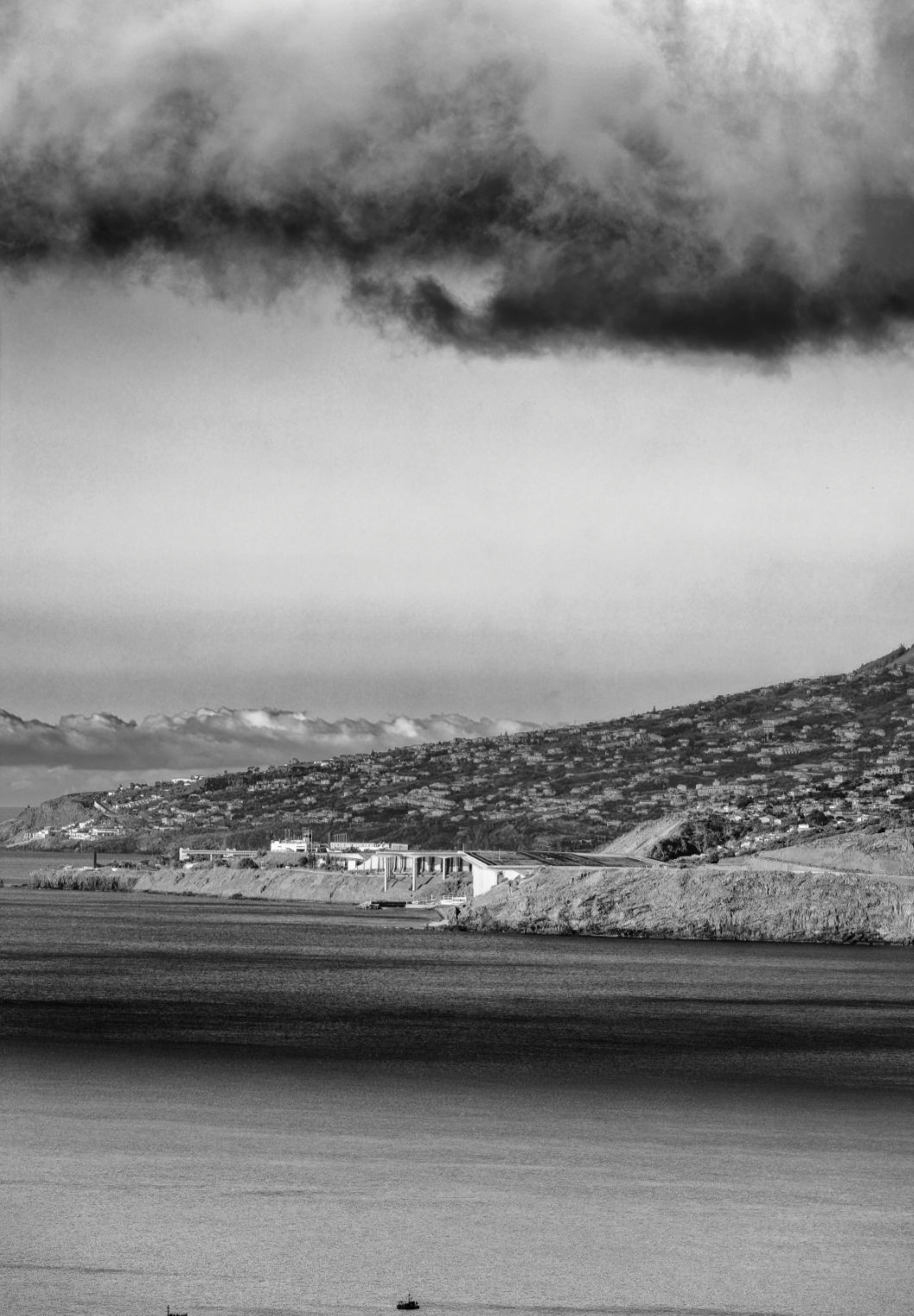 Schwarzweiß Photo von Madeira. Im oberen Bereich dramatische Wolken. Im unteren Bereich das Meer mit einem kleinen Boot. Im mittleren Bereich ist ein Teil der Insel zu sehen. Die Landebahn des Flughafen ist etwa in der Mitte. 