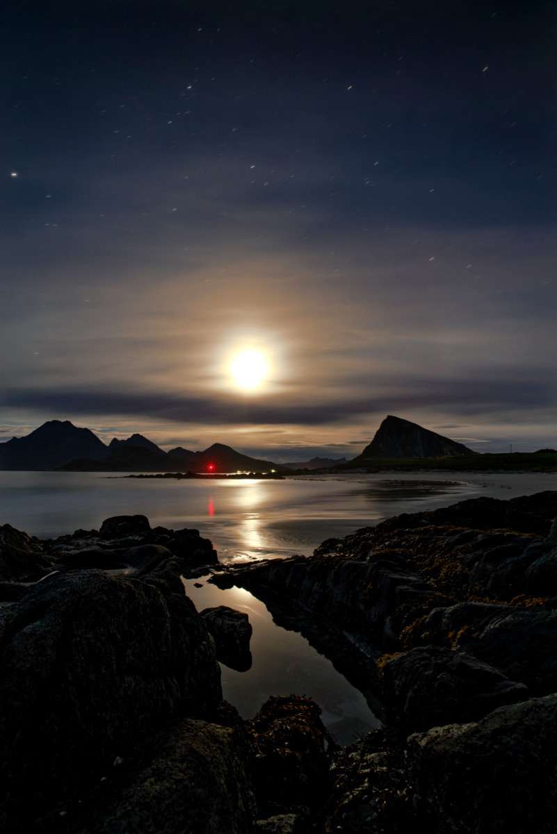 Dunkle Küsten Szene. Im Vordergrund zwei Felsen zwischen denen sich ein Pool bildet. Im Mittelgrund das Meer mit etwas Strand am rechten Rand sowie die in den Hintergrund übergehenden Berge. Der Mond scheint weich durch die Schleierwolken. Am Nachthimmel sind ein paar Sterne zu sehen. 