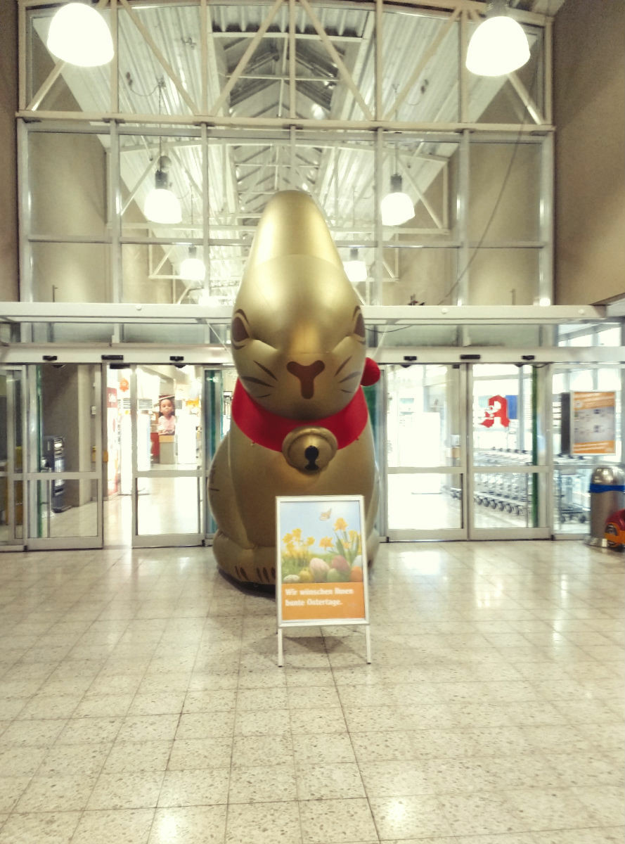 Am Ausgang eines Großmarkt steht ein doppelt Mannshoher aufblasbarer Lindt Schokohase und blickt einen an. Vor ihm steht ein aufsteller mit einem 'frohe Ostern ' Aufdruck. 