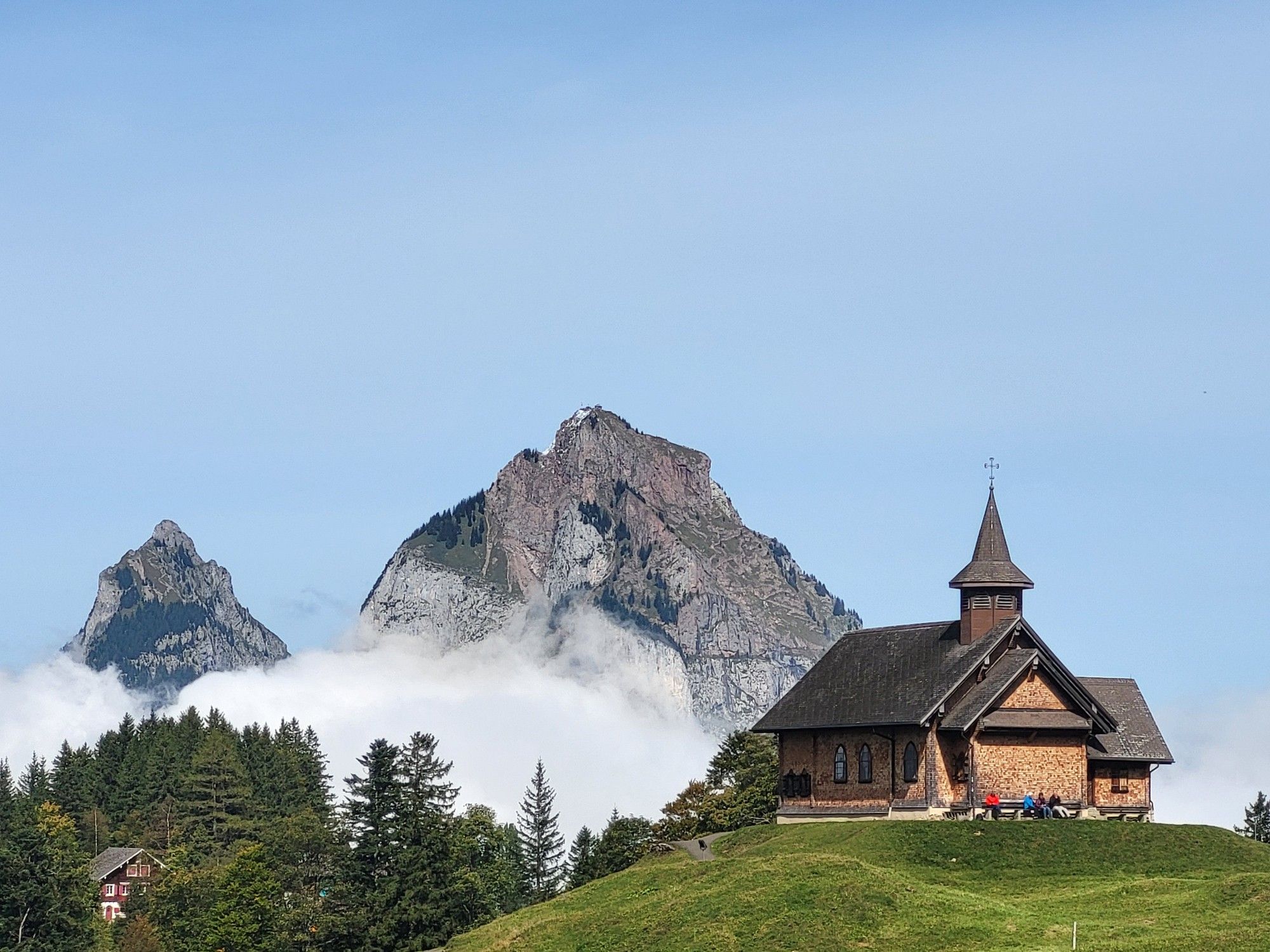 Church on a green hill, clouds contrasting in the background, with two rocky mountain peaks not far behind the clouds.