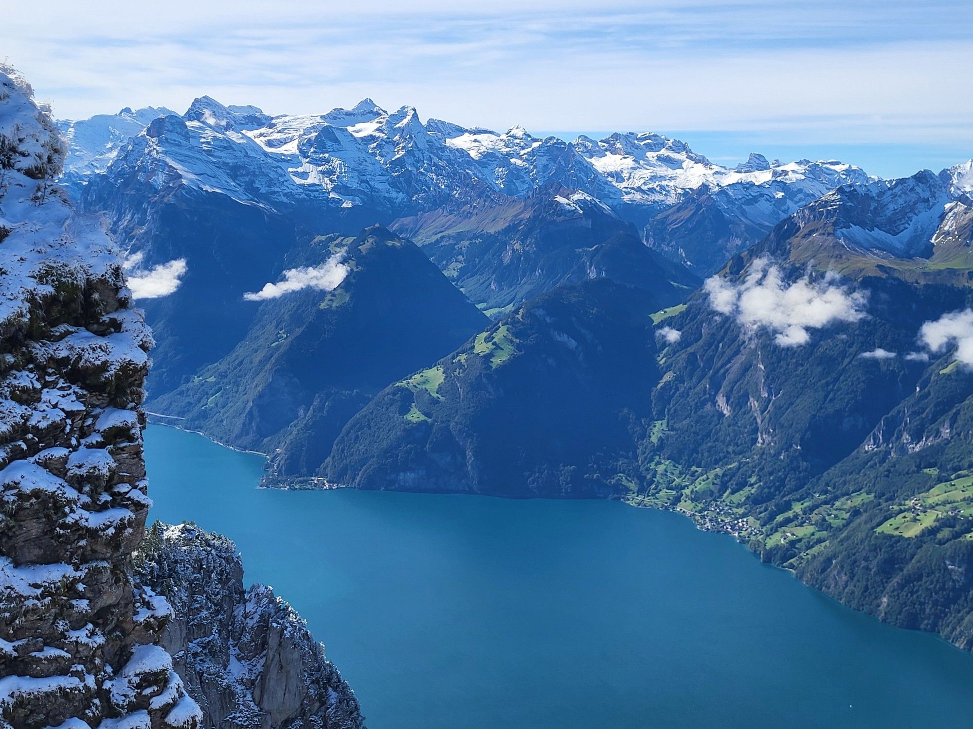 Looking down at a lake surrounded by very steep dramatic mountains, snow topped peaks. A few clouds float in front of the mountains. There's a snowy steep rocky precipice close to the photographer framing the lake on the left.