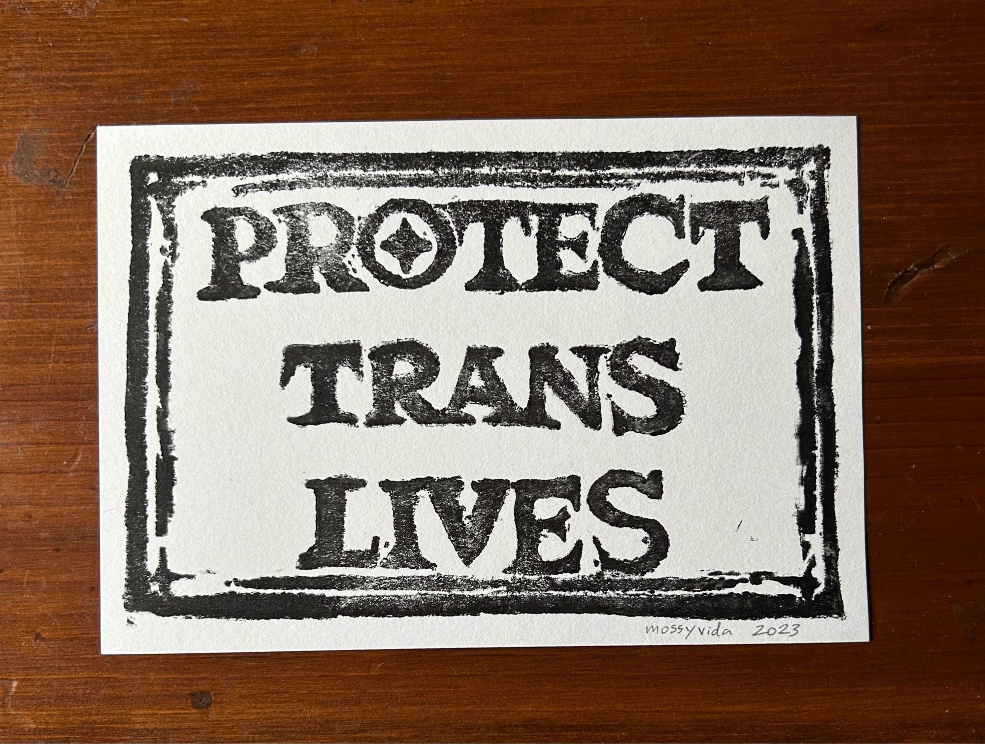 A linocut print pictured on a dark wooden table. The paper is white with black stamped letters that reads: PROTECT TRANS LIVES. It’s signed “mossyvida 2023” on the bottom right corner in pencil.