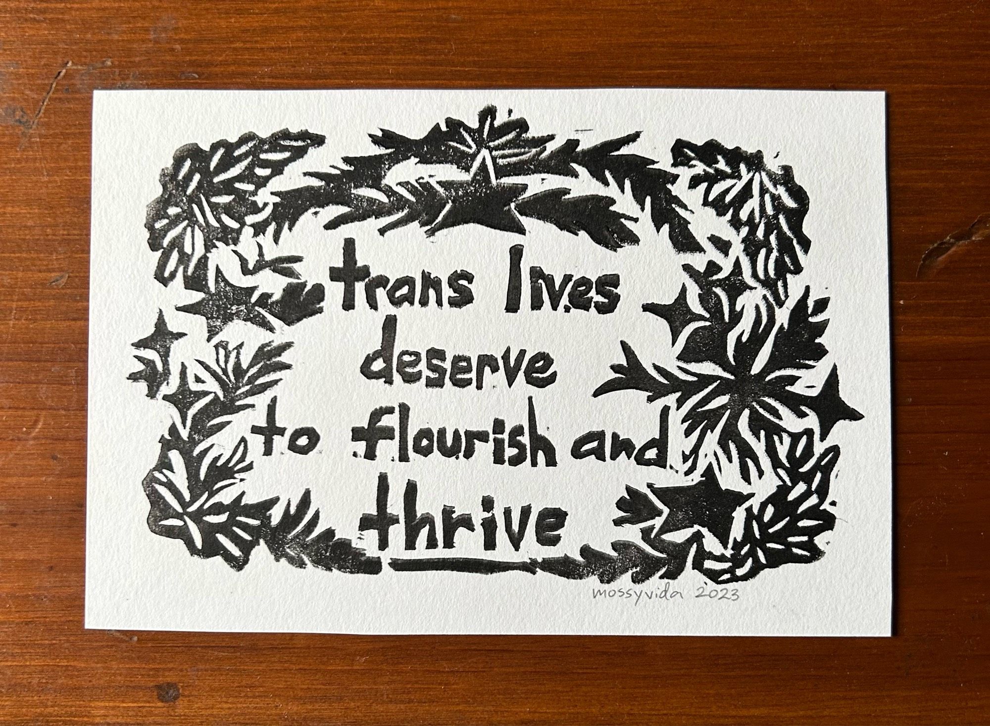 A linocut print pictured on a dark wooden table. The paper is white with black stamped letters that reads: “trans lives deserve to flourish and thrive”with stars and foliage surrounding the text. It’s signed “mossyvida 2023” on the bottom right corner in pencil.