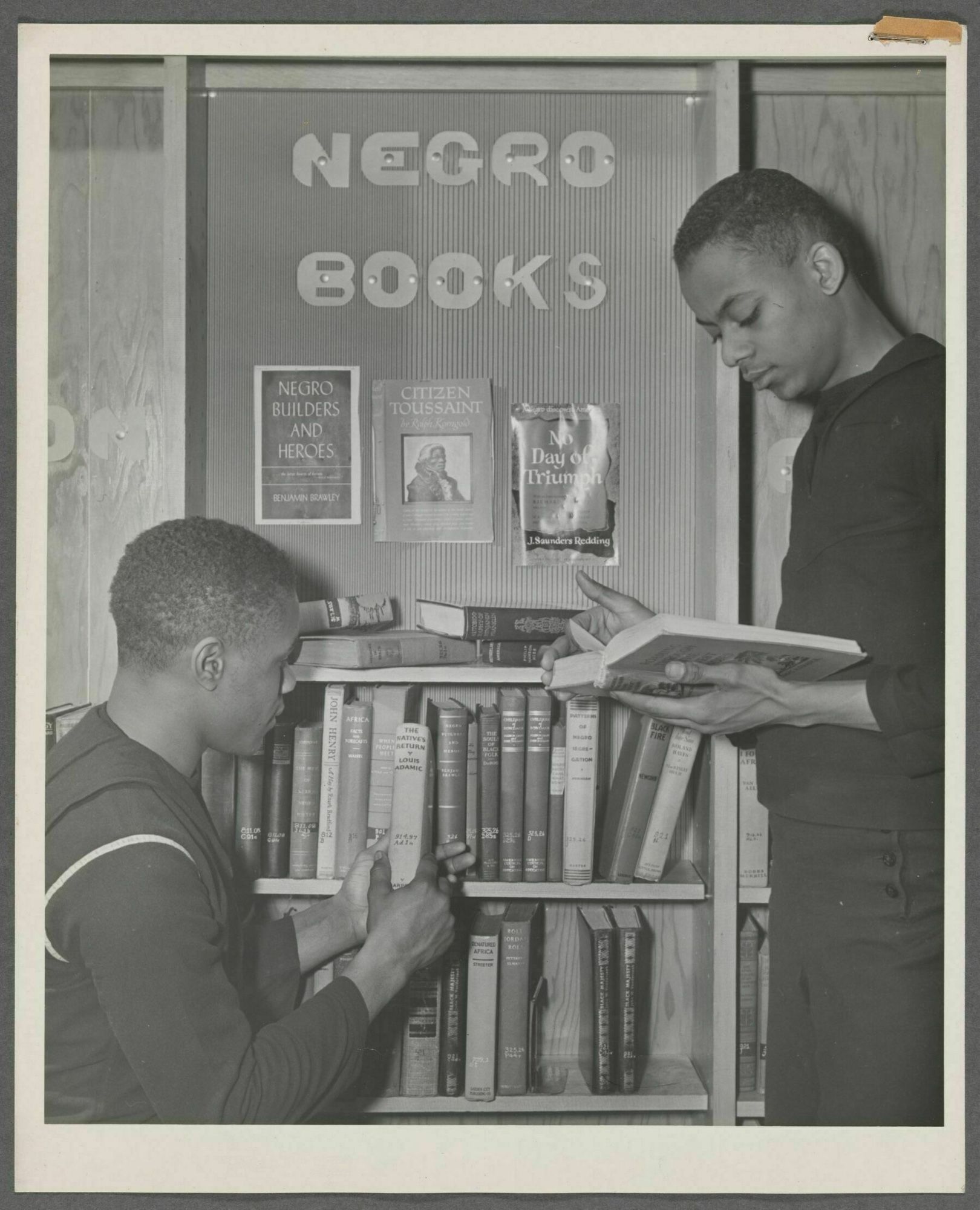 Black and white photo of two sailors browsing books at a small library section advertising “Negro Books”