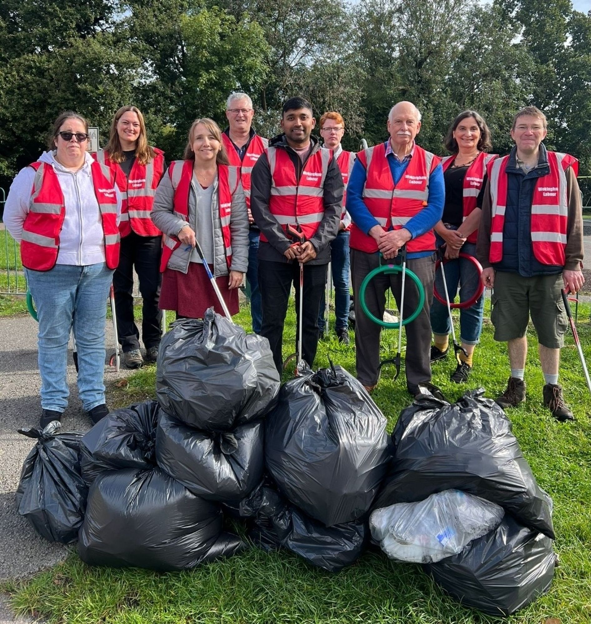 Photo shows a mixed group of residents, councillors and Labour volunteers standing in two lines behind a small wall of black plastic bags,  filled with litter. One of the bags is clear and contains recyclable litter.

Everyone is wearing Wokingham Labour red high visibility vests. Some of the group are holding litter picking sticks, some are holding plastic hoop.

The bags are on grass, about 30 metres behind the group are some trees.