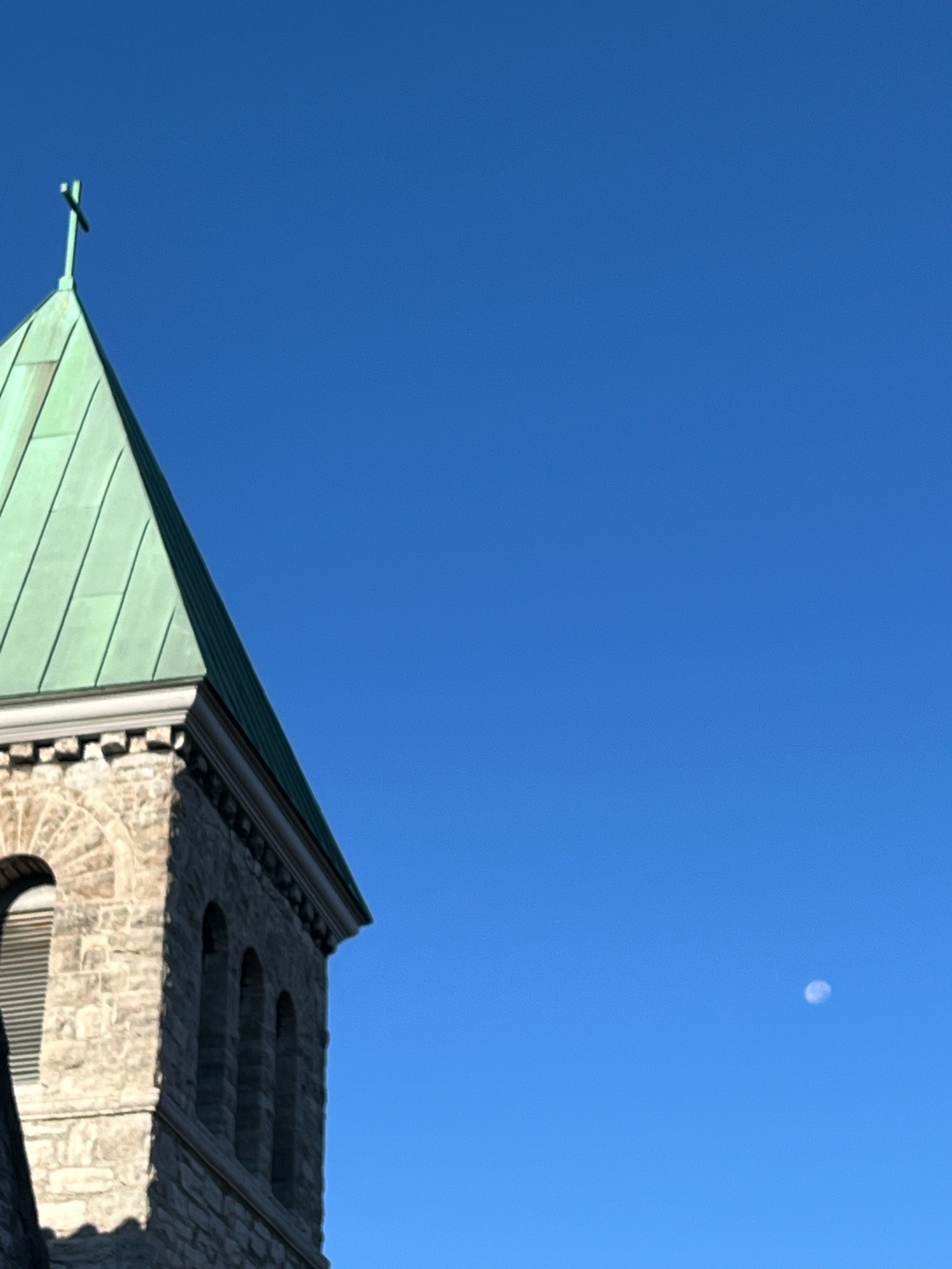 Most of a moon visible in blue sky beside the limestone bell tower of a Richardsonian Romanesque Episcopalian church in Buffalo, New York 