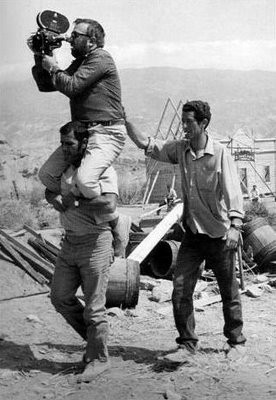 En la imagen se ve a Sergio Leone sobre los hombros de un operario mientras sostiene una cámara de cine. Tras él, otro operario lo sujeta por la espalda para que no se desequilibre.