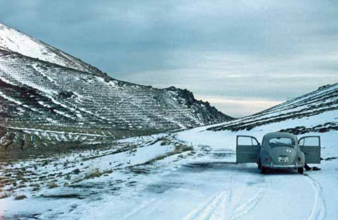 La imagen muestra un paisaje de montaña nevado que corresponde al Puerto de la Ragua en Sierra Nevada. Hacia el margen derecho de la imagen, aparece detenido un Volkswagen Escarabajo del que se aprecia su parte trasera.