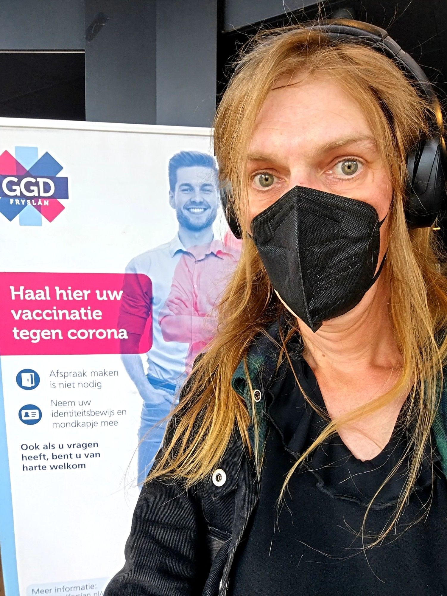 Ik bezweet en met m'n haar door de war voor het bord van de GGD waarop haal hier uw vaccinatie tegen corona staat. Afspraak maken niet nodig