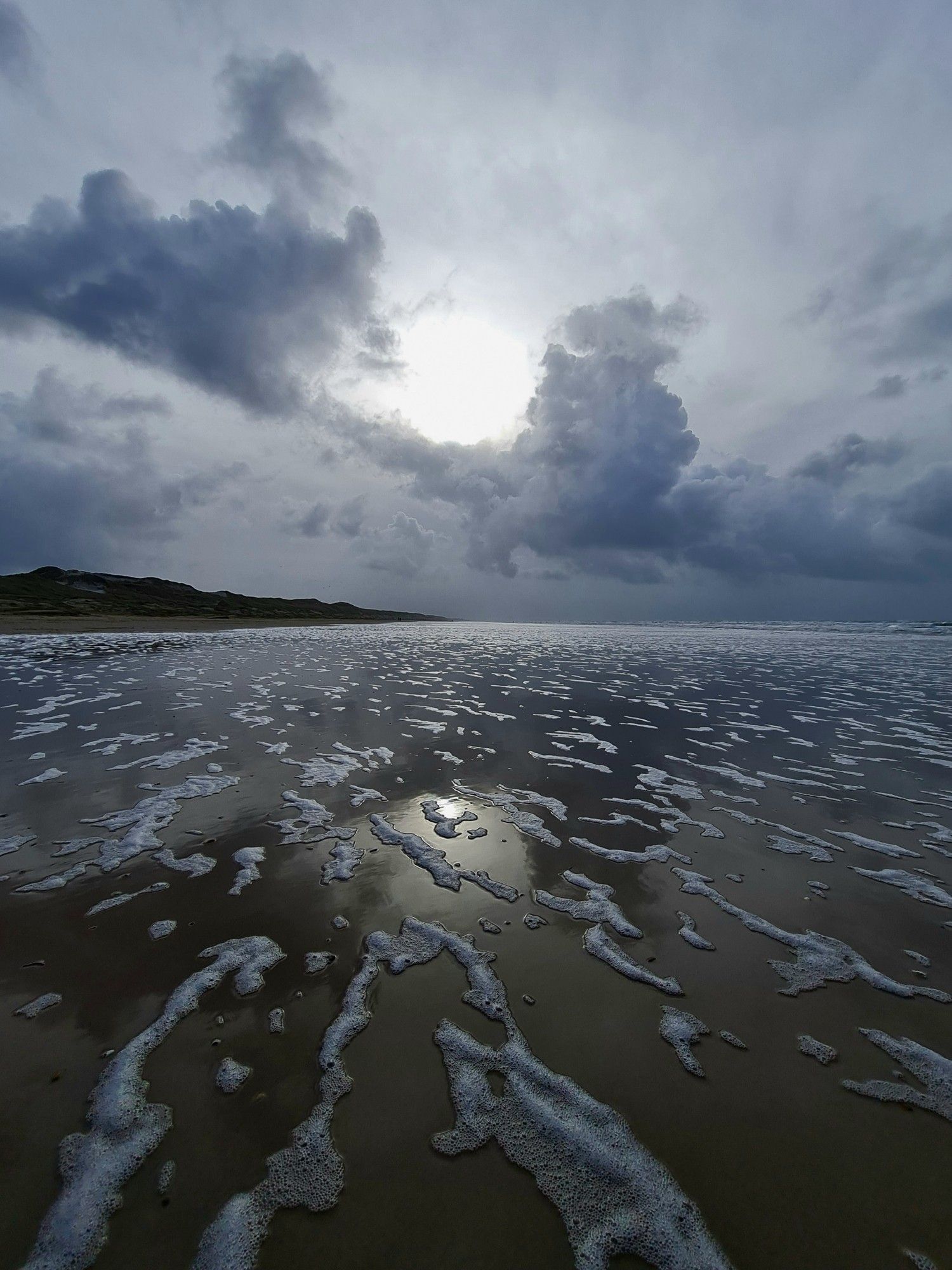 A photo of a wet beach, a thin sliver of dunes and large clouds