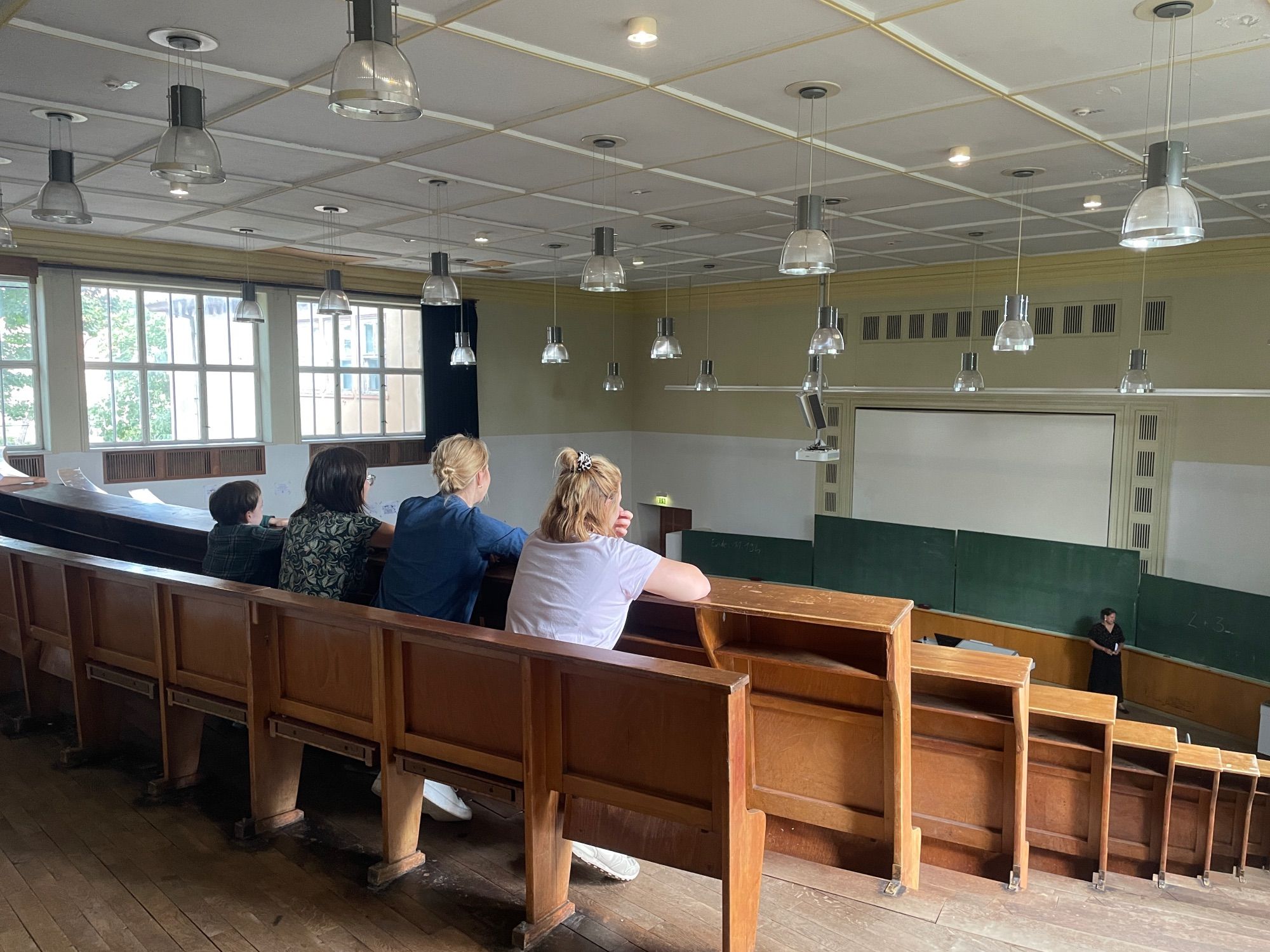 Von hinten sind drei Erwachsene und ein Kind zu sehen, die in der obersten Stuhlreihe des Hörsaals sitzen, dessen großer Raum mit vielen Stuhlreihen, Lampen und der Tafel im unteren Bereich zu sehen ist.