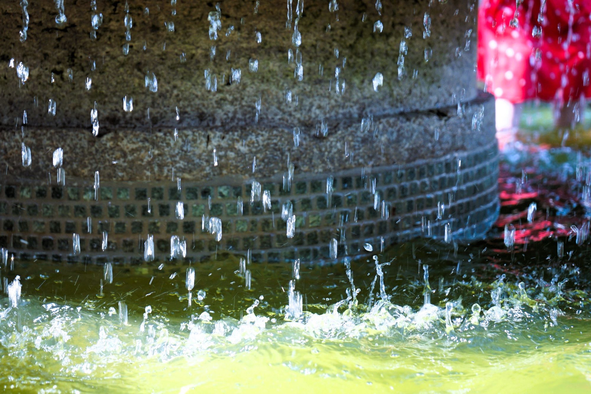 Das Bild zeigt Wassertropfen und verschwommen ein Kind, das mit den Beinen im Wasser steht.