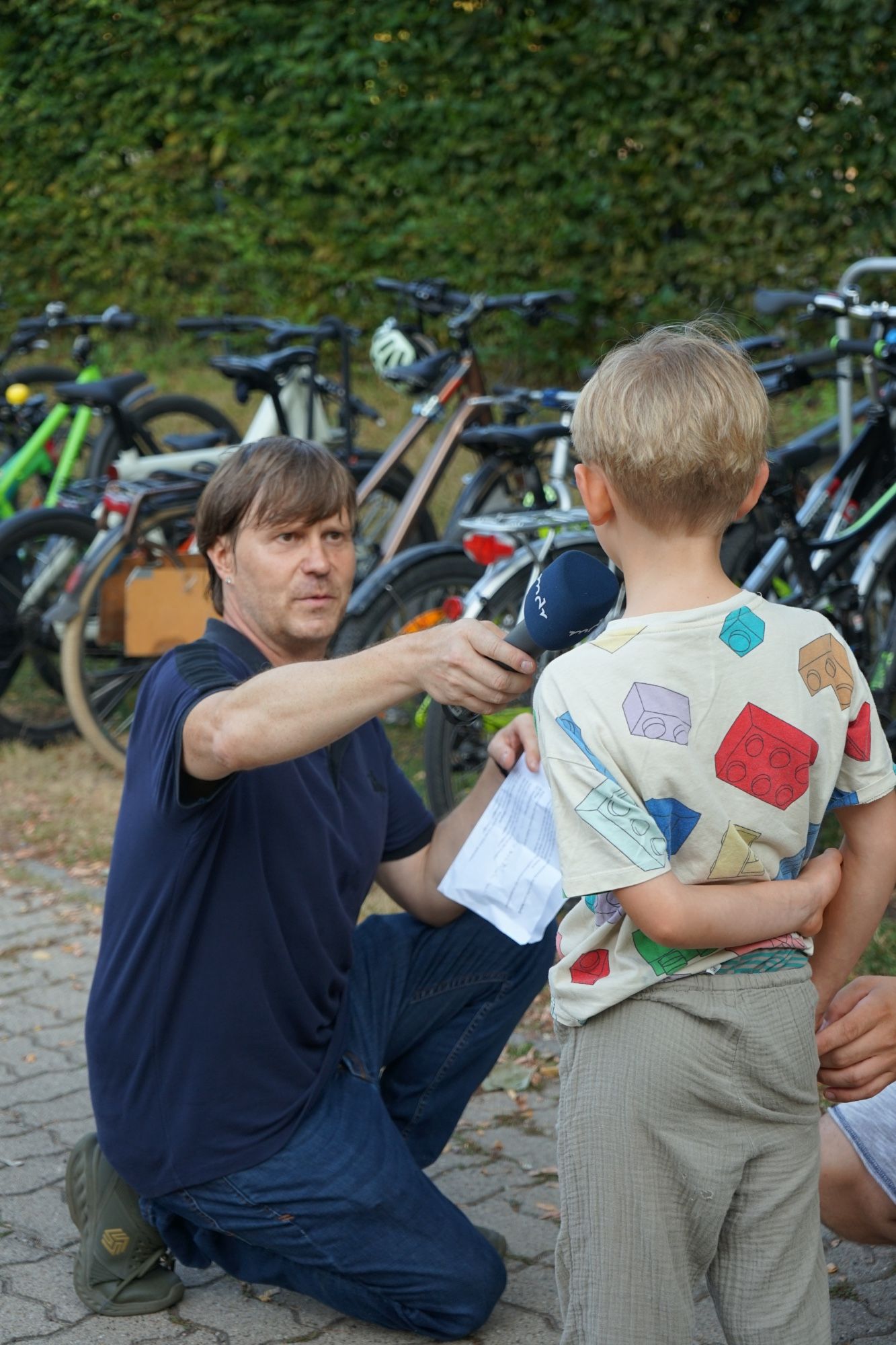 Vor einem Fahrradständer voller Fahrräder kniet ein Mann in dunkelblauer Kleidung und hält einem Kind in heller Kleidung, das von hinten zu sehen ist, ein Mikrofon mit der Aufschrift mdr hin.