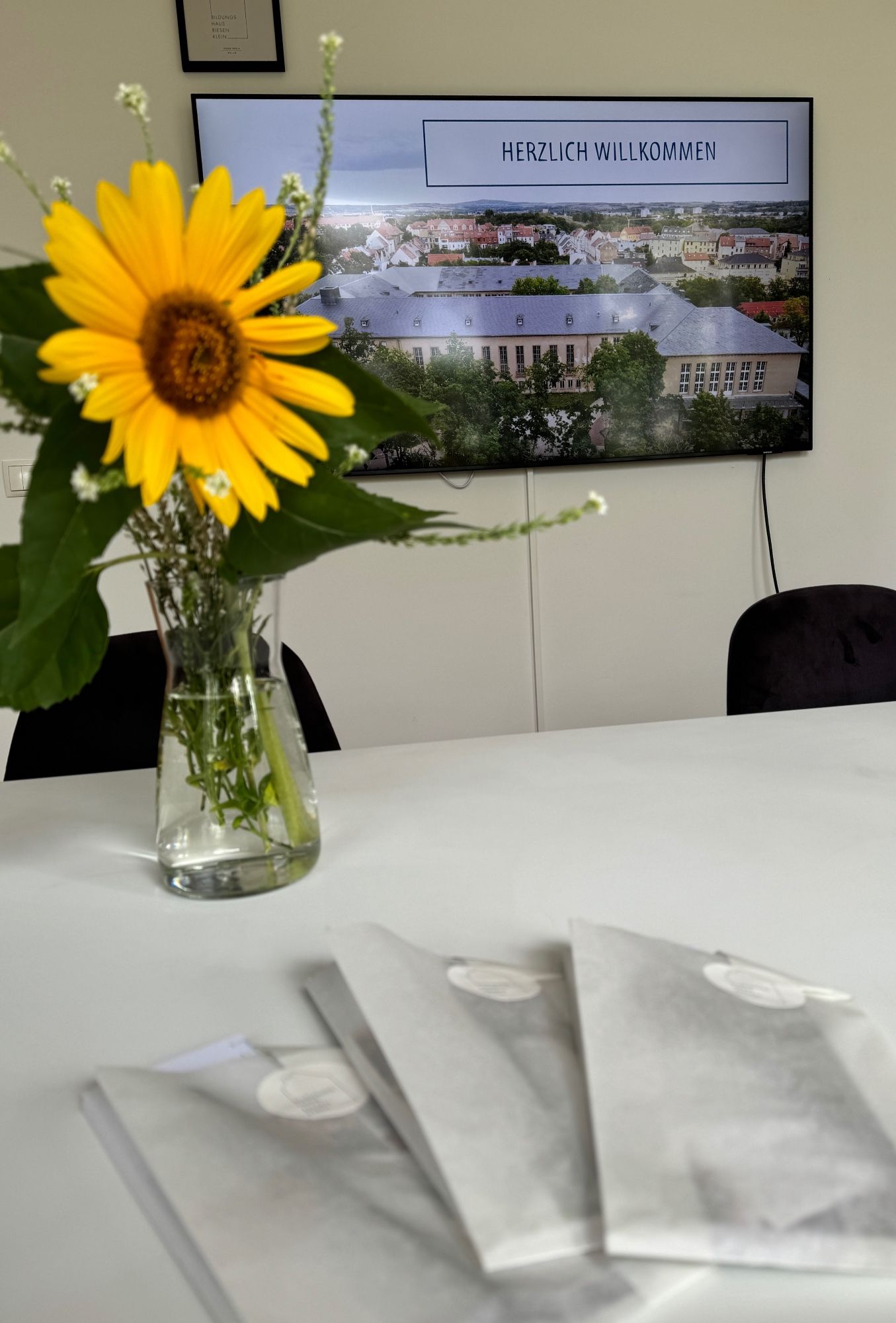 Zu sehen ist ein Präsentationsbildschirm mit der Aufschrift „Herzlich willkommen“, davor eine Sonnenblume und drei Geschenktüten auf dem Tisch.