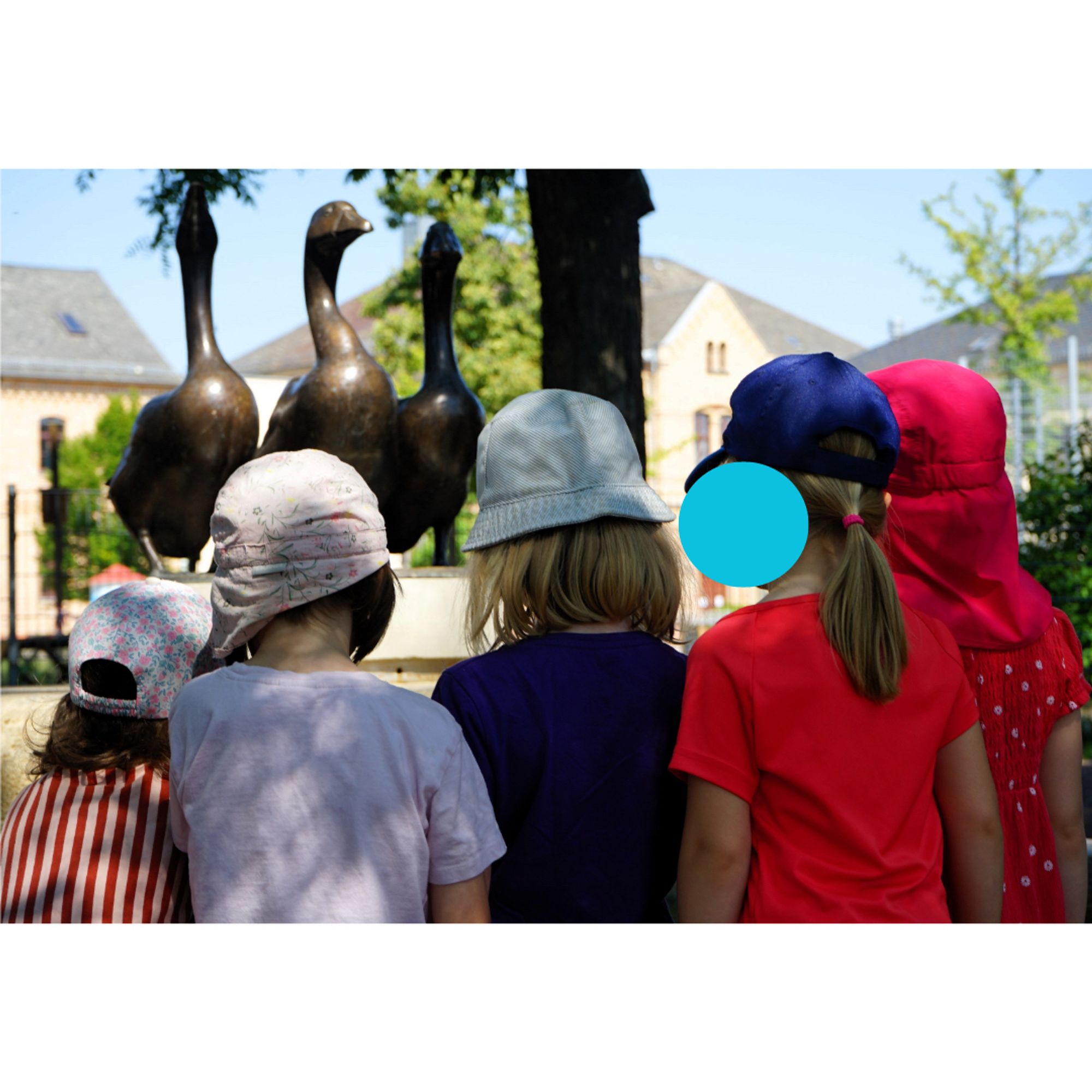 Das Bild zeigt 5 Kinder in farbigen T-Shirts und Sonnenhüten von hinten, die vor einem Brunnen mit drei Gänse-Skulpturen stehen.