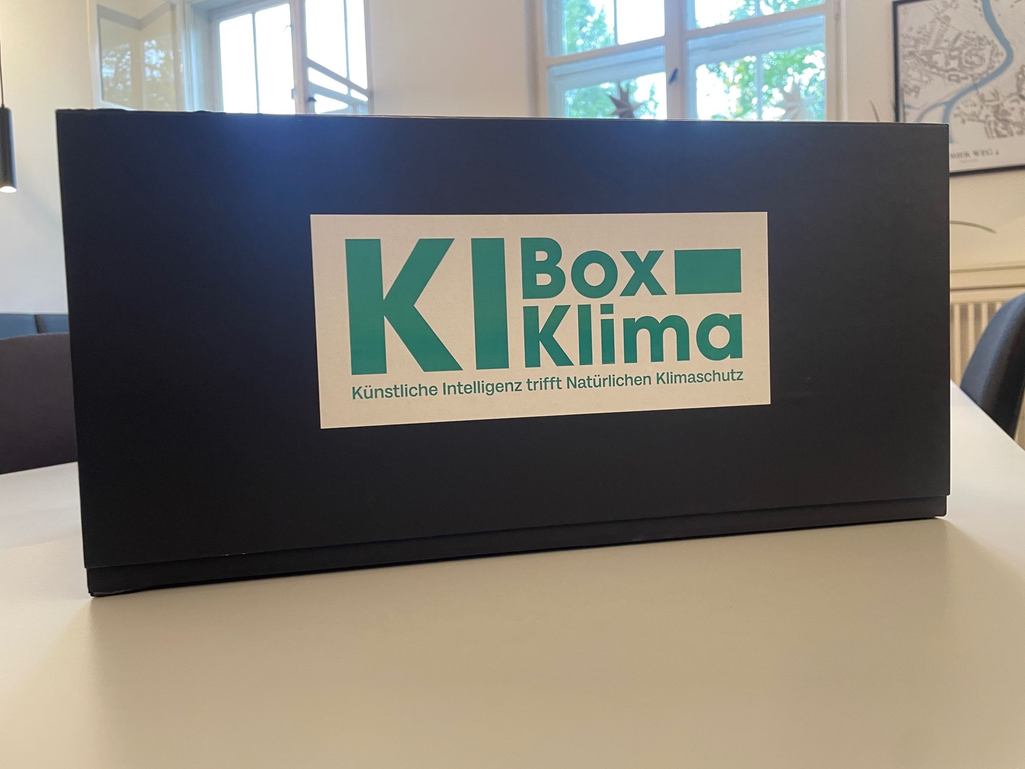 Das Bild zeigt eine große schwarze Kiste, die auf einem Tisch steht, mit der Aufschrift „KI Box Klima – Künstliche Intelligenz trifft Natürlichen Klimaschutz“.