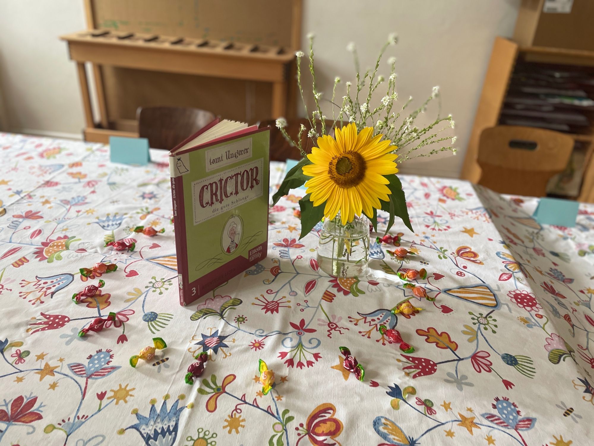 Auf einer bunt geblümten Tischdecke steht eine Vase mit einer großen gelben Sonnenblume, daneben das Buch „Crictor, die gute Schlange“ und umrundet von bunten Bonbons.