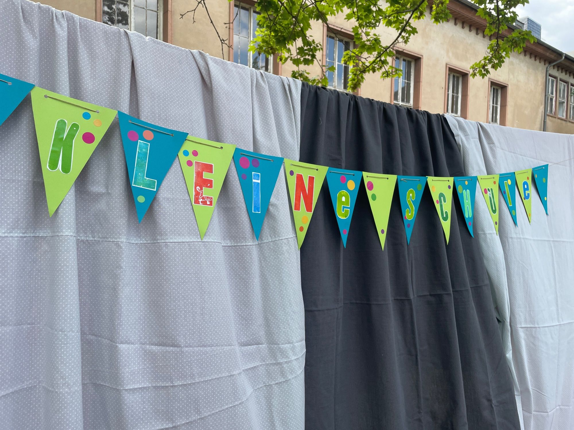 Vor einem grau/grau-gepunkteten Vorhang hängt eine grün-blaue Wimpelkette, auf der in großen bunten Buchstaben „KLEINE SCHULE“ steht.