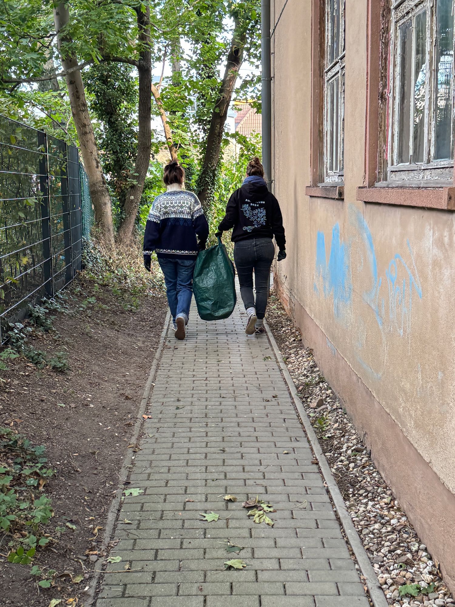 Das Bild zeigt auf dem Weg neben dem Haus zwei Frauen von hinten, die gemeinsam einen großen Gartenabfallbehälter zwischen sich tragen.