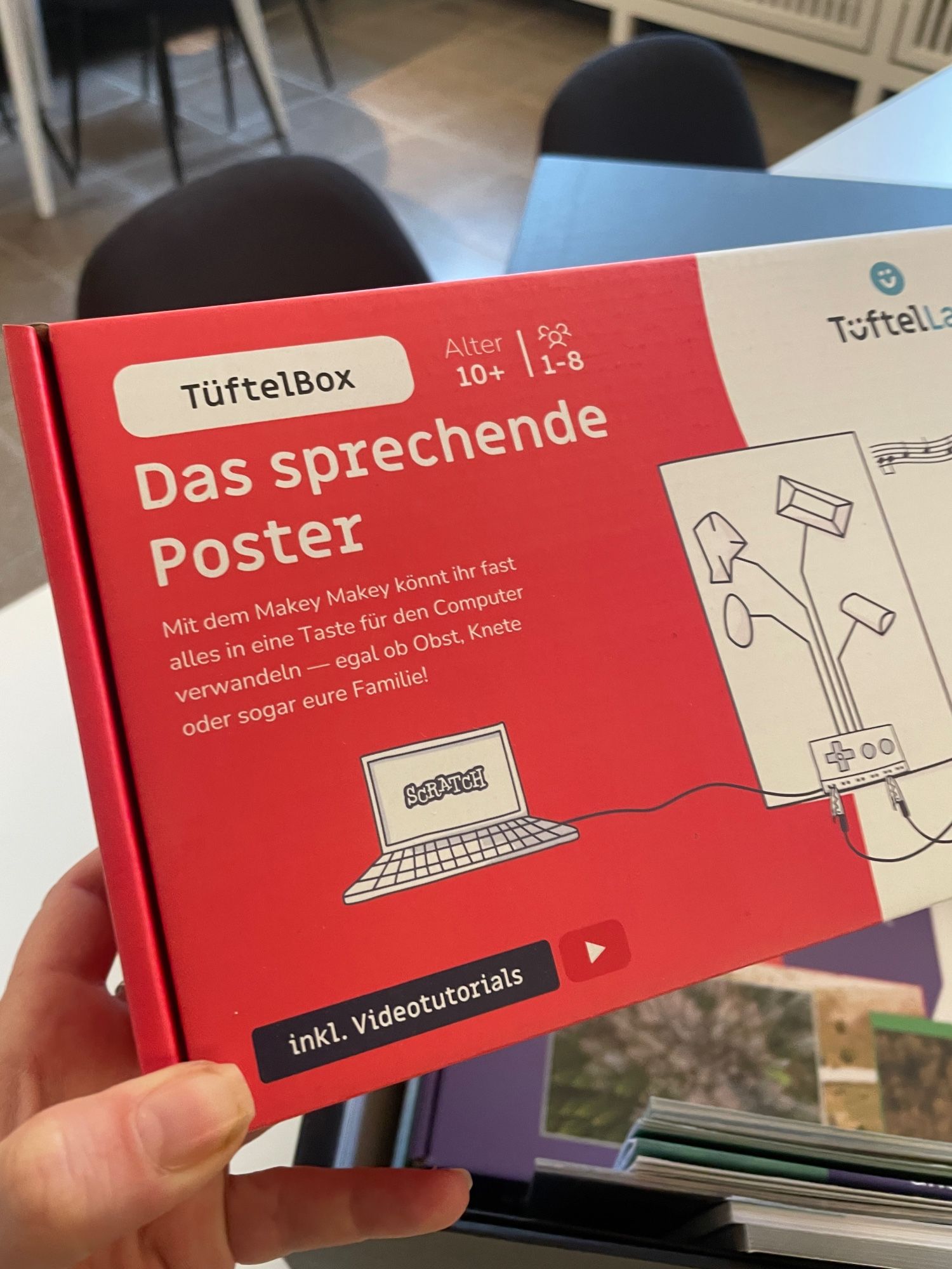 Zu sehen ist eine Hand, die eine Packung mit der Aufschrift „TüftelBox Das sprechende Poster“ aus der Kiste nimmt.